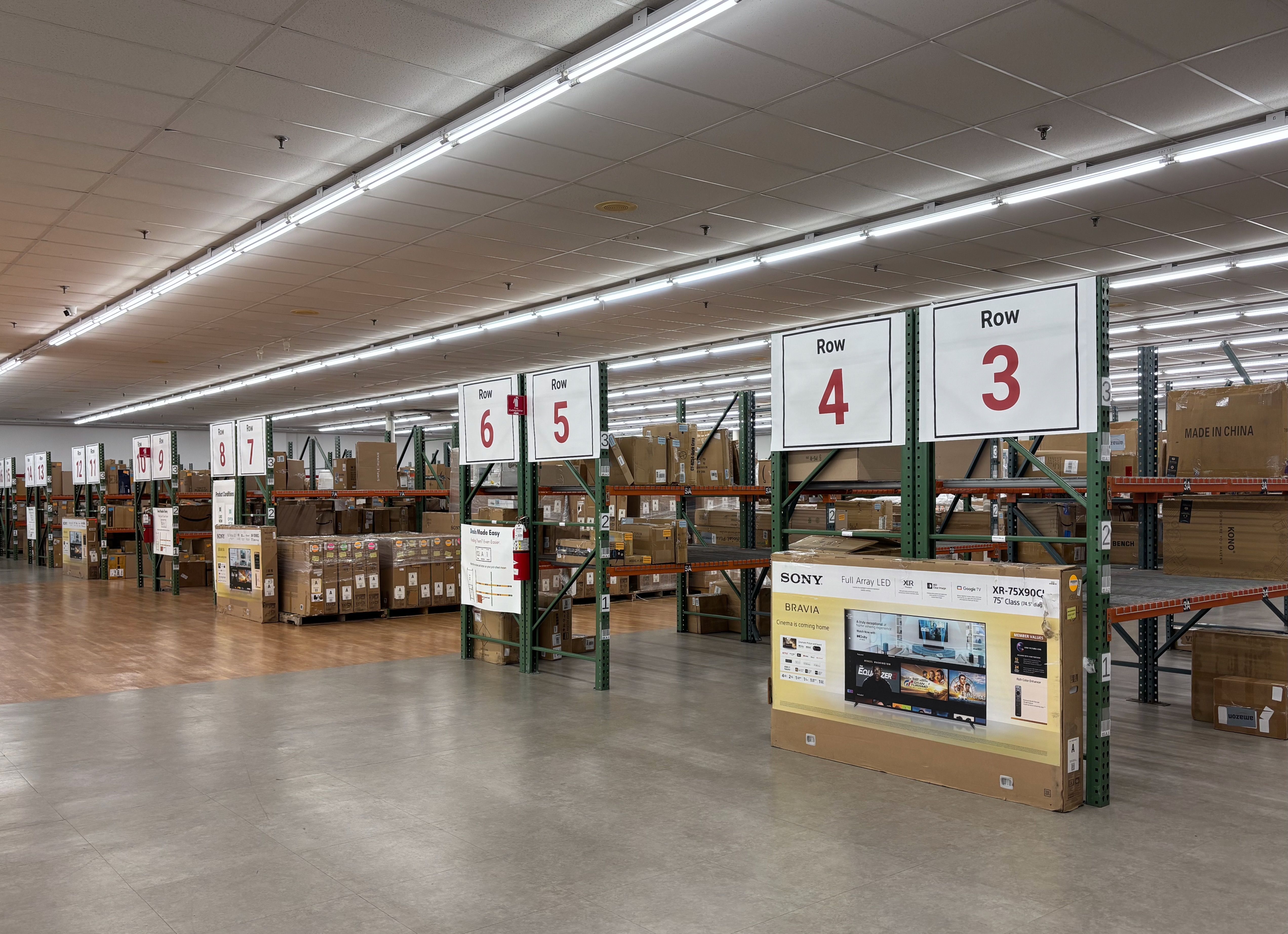 Large warehouse with numbered storage rows from 3 to 13, filled with assorted large cardboard boxes and appliances, fluorescent ceiling lights, and concrete floors.