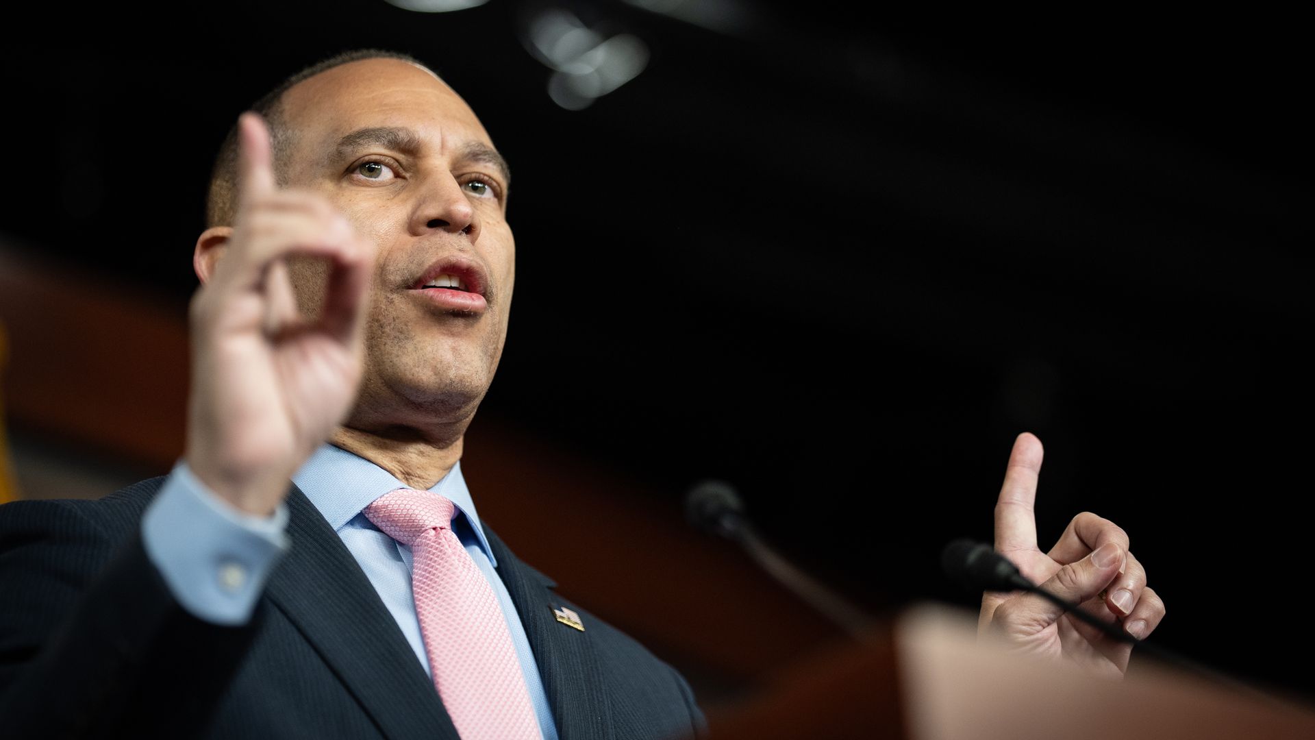 Man in dark suit, light blue shirt, and pink tie speaking and gesturing with raised index fingers at a podium with microphones in a dark room.