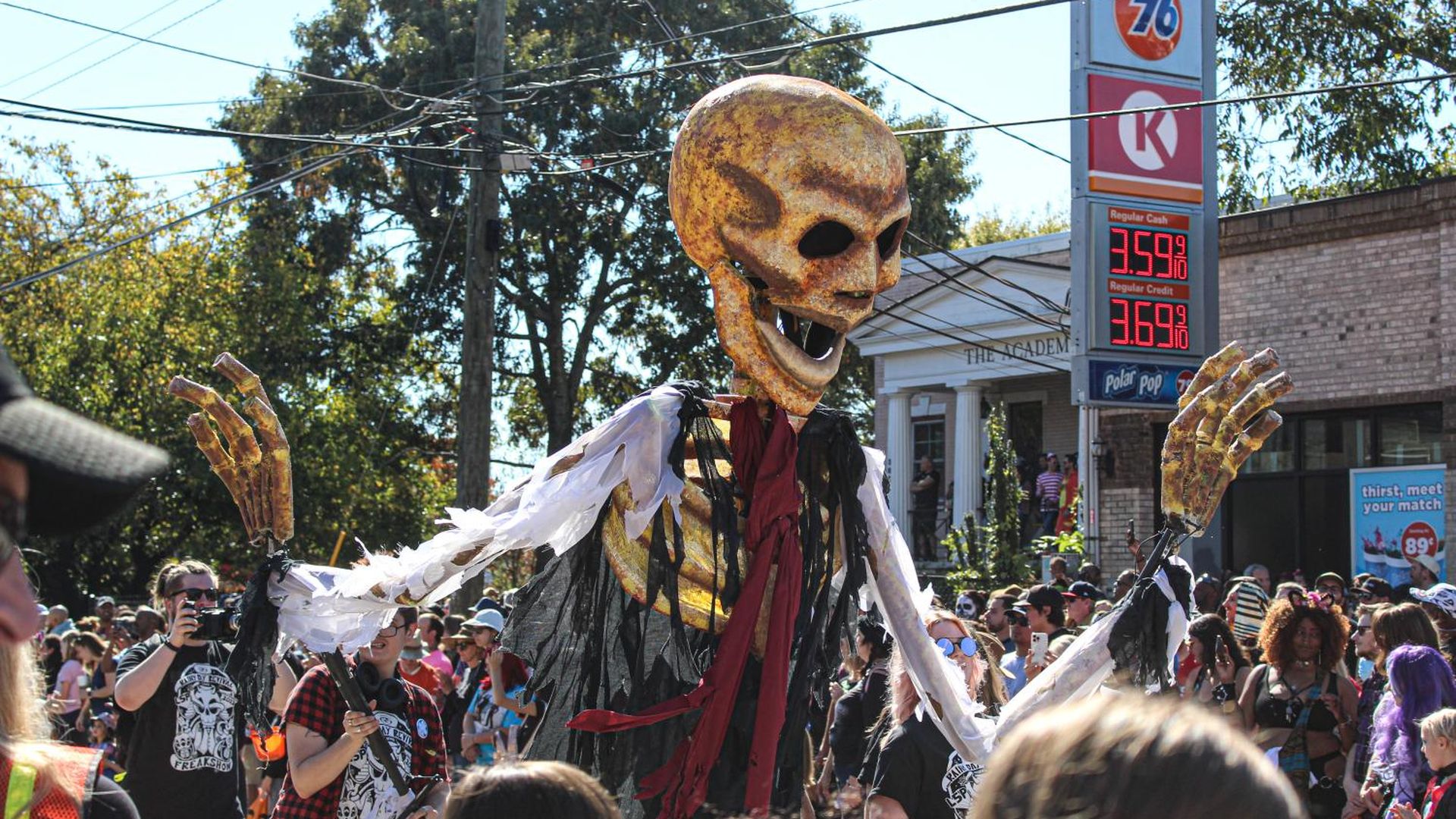 A large puppet of a skeleton with a large skull covered in cobwebs waves at a parade crowd
