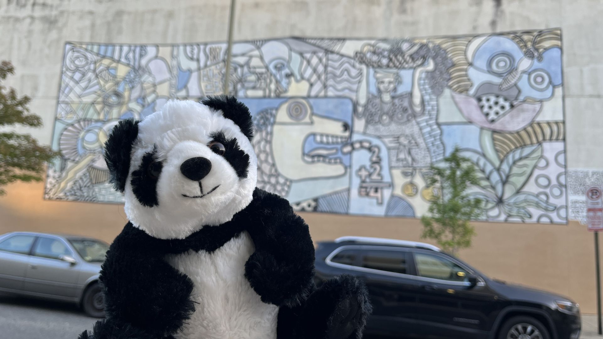 A hand holds up a stuffed animal panda in front of a large mural in Adams Morgan.