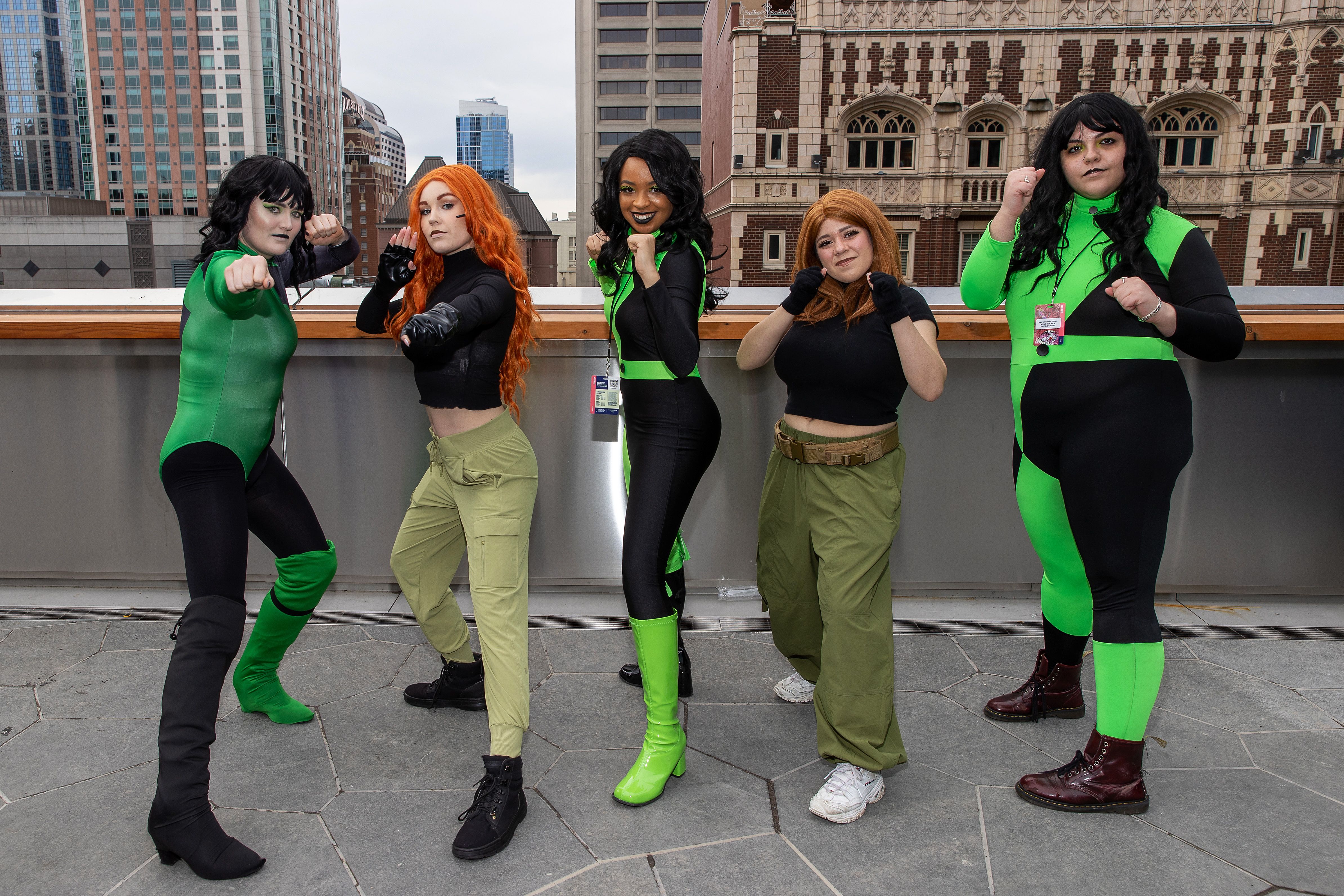 Five women in black and green cosplay gear stand on a rooftop with their arms up in fighting poses.