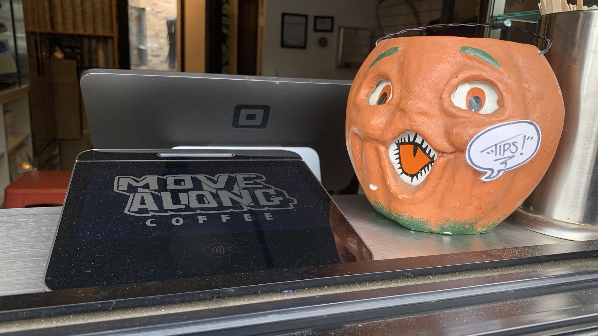 Close-up of a coffee shop counter showing a payment terminal with "MOVE ALONG COFFEE" logo and an orange pumpkin-shaped tip jar with a cartoon face and a speech bubble saying "TIPS!".