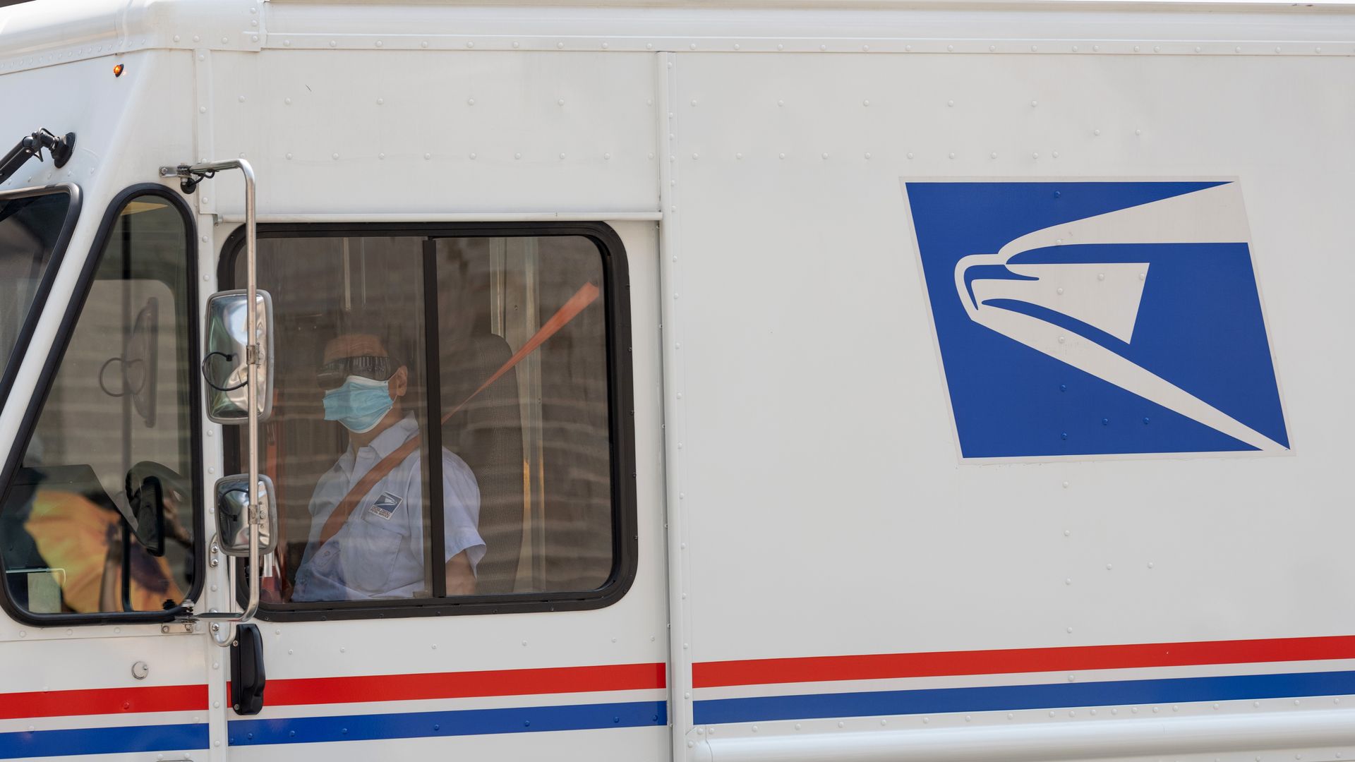 A driver wearing a face mask drives a USPS mail truck 