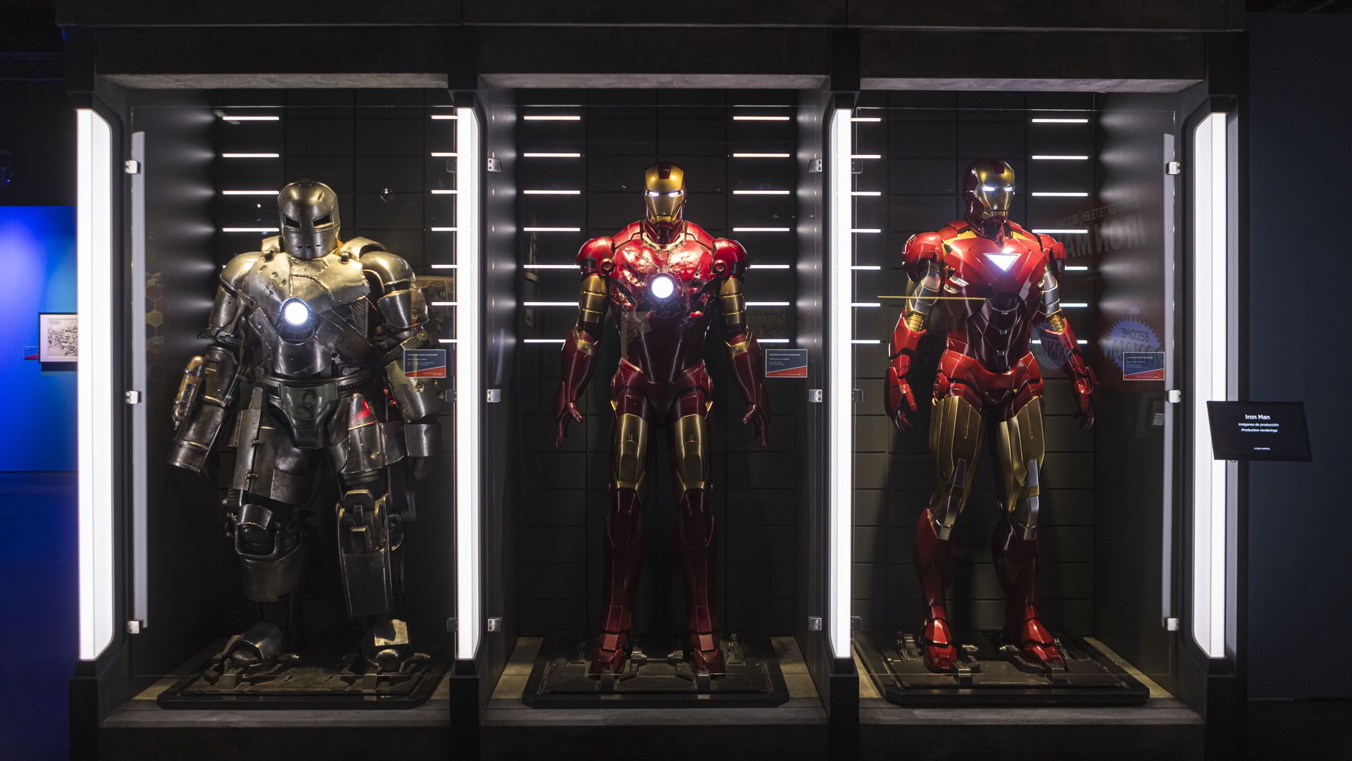 Three Iron Man suits on display in glass cases against a dark backdrop: left a silver War Machine armor, center a red-and-gold Iron Man suit, right another red-gold suit with a glowing chest reactor.
