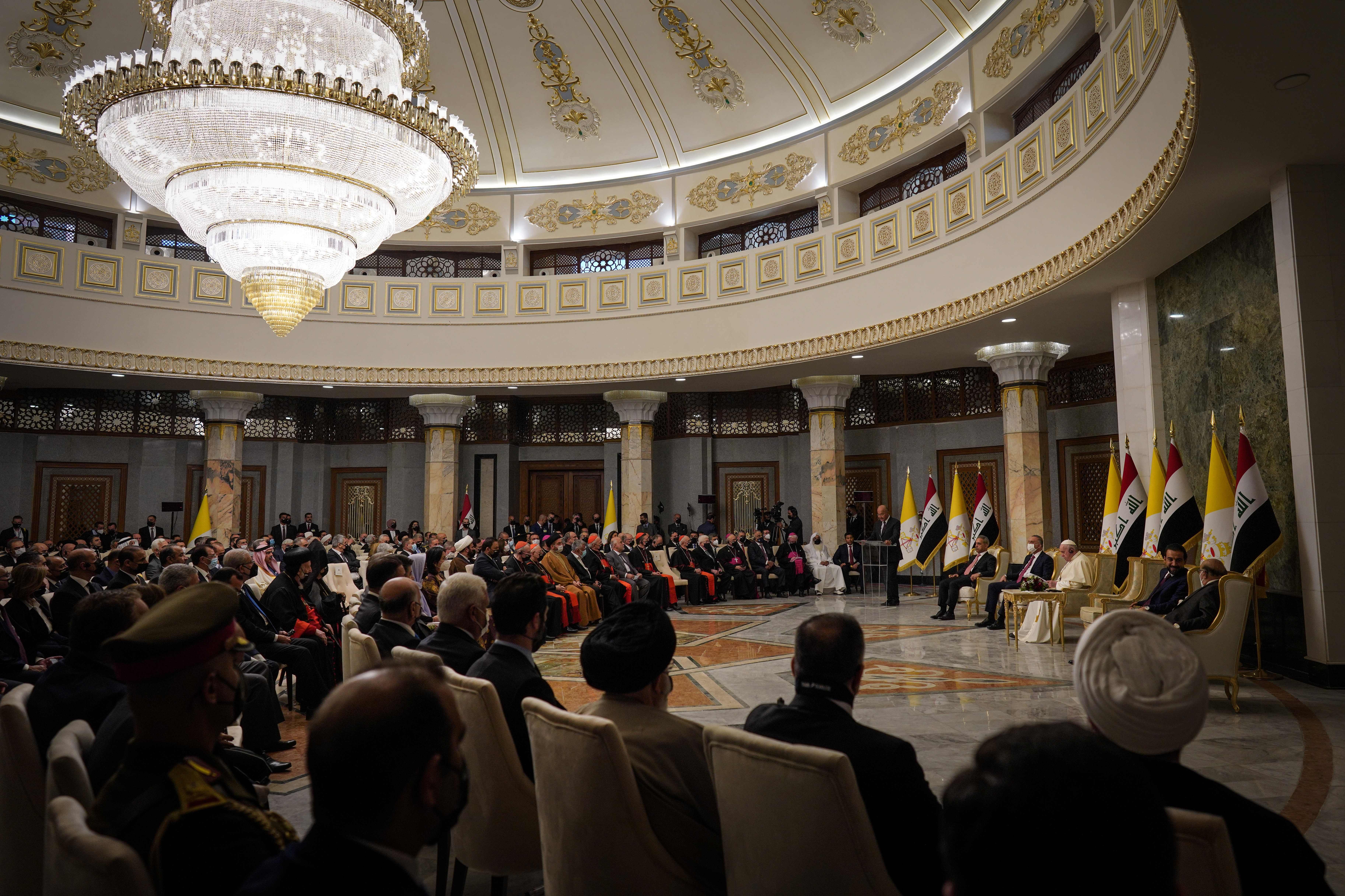 picture of the Iraq presidential palace, with a large crowd of people sitting on chairs looking at the Pope as he delivers a speech.
