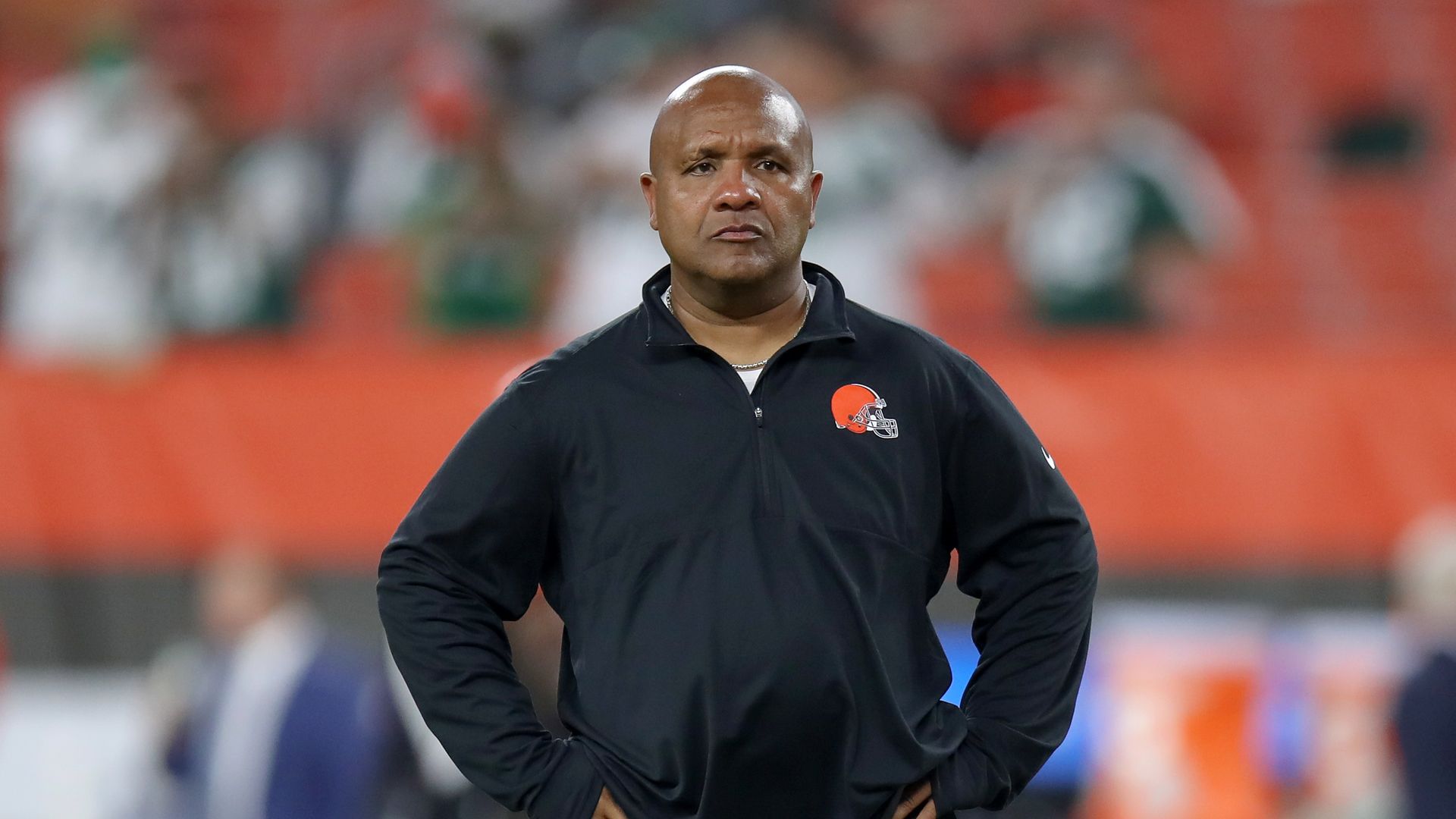 A bald man wearing a black zip-up jacket with an orange football helmet logo stands on a field, hands on hips, with a blurred orange stadium backdrop.