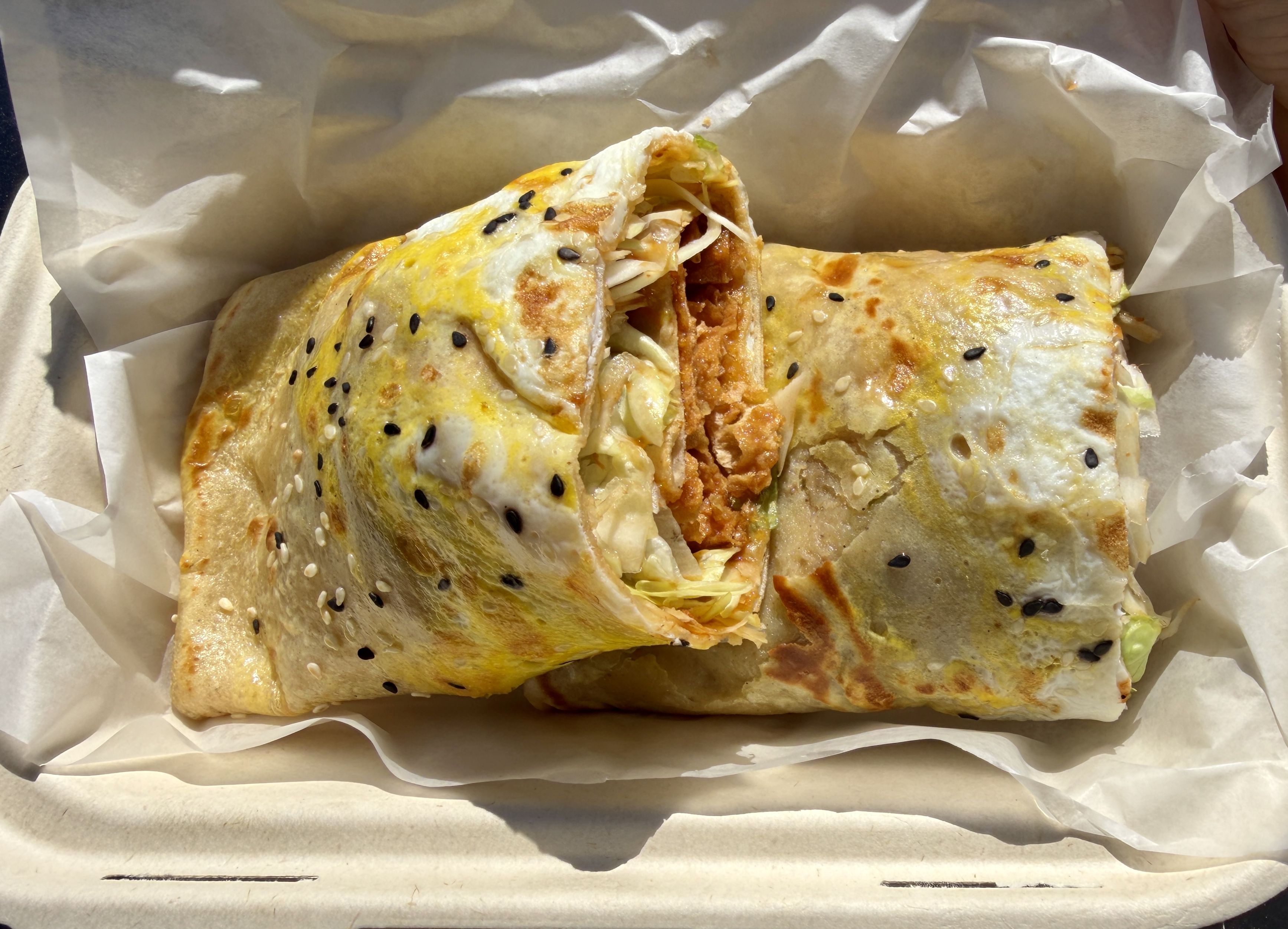 Sesame-seeded flatbread wrap, toasted and split open to reveal crispy orange chicken, shredded lettuce, and melted cheese inside on parchment.