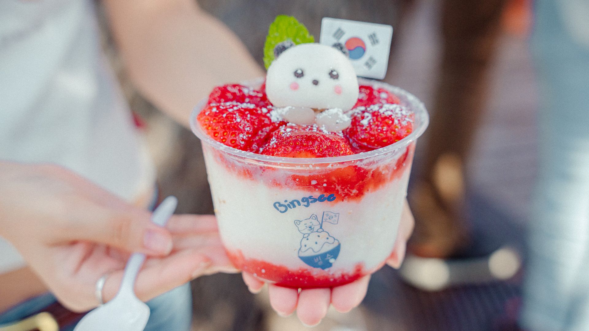 Cup of strawberry bingsu topped with a cute white marshmallow panda, pink cheeks, and a green leaf; a small card with the Korean flag sits behind as red strawberry sauce surrounds the dessert.