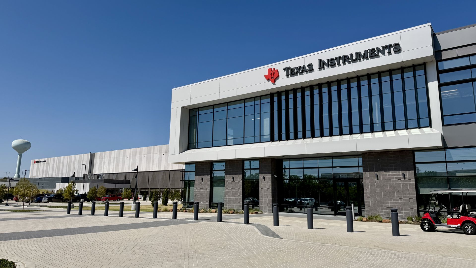 Modern Texas Instruments office building with a white facade, large glass windows, and a red TI logo. Bright blue sky, a paved lot with bollards, and a red golf cart on the right.