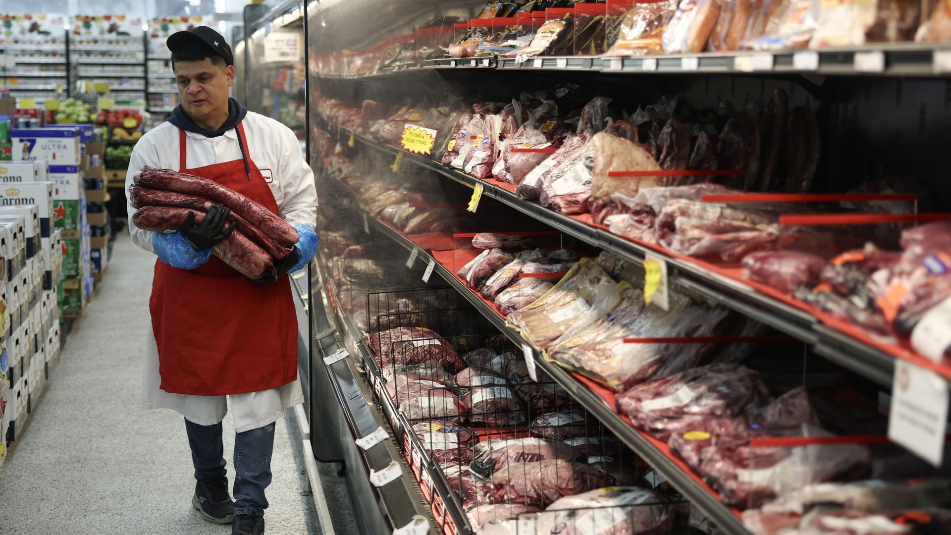 A man in a white coat and red apron holds large pieces of raw meat inside a supermarket meat aisle with shelves stocked with various packaged meats.