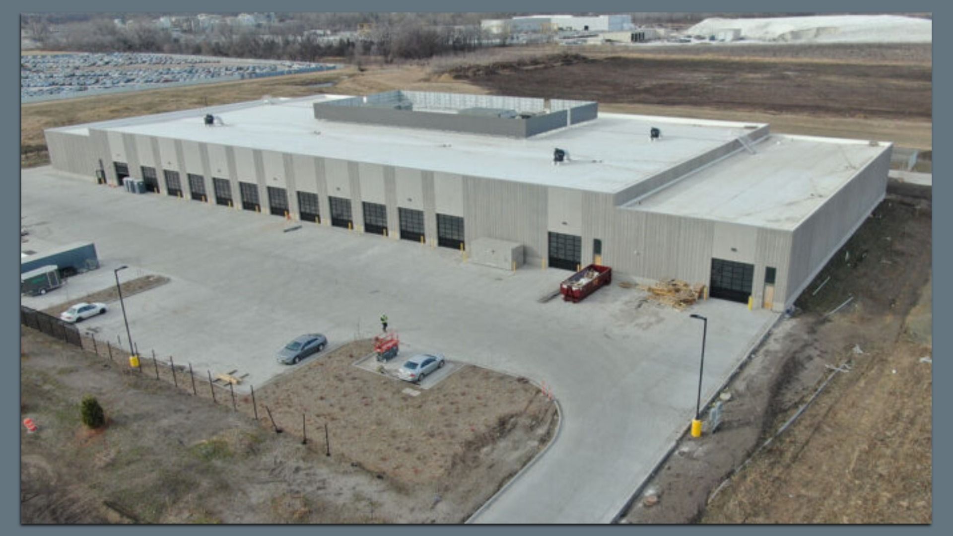 A large gray industrial warehouse with several loading bays and a flat white roof. A red truck sits by pallets near the loading area, as a curved driveway circles the building.