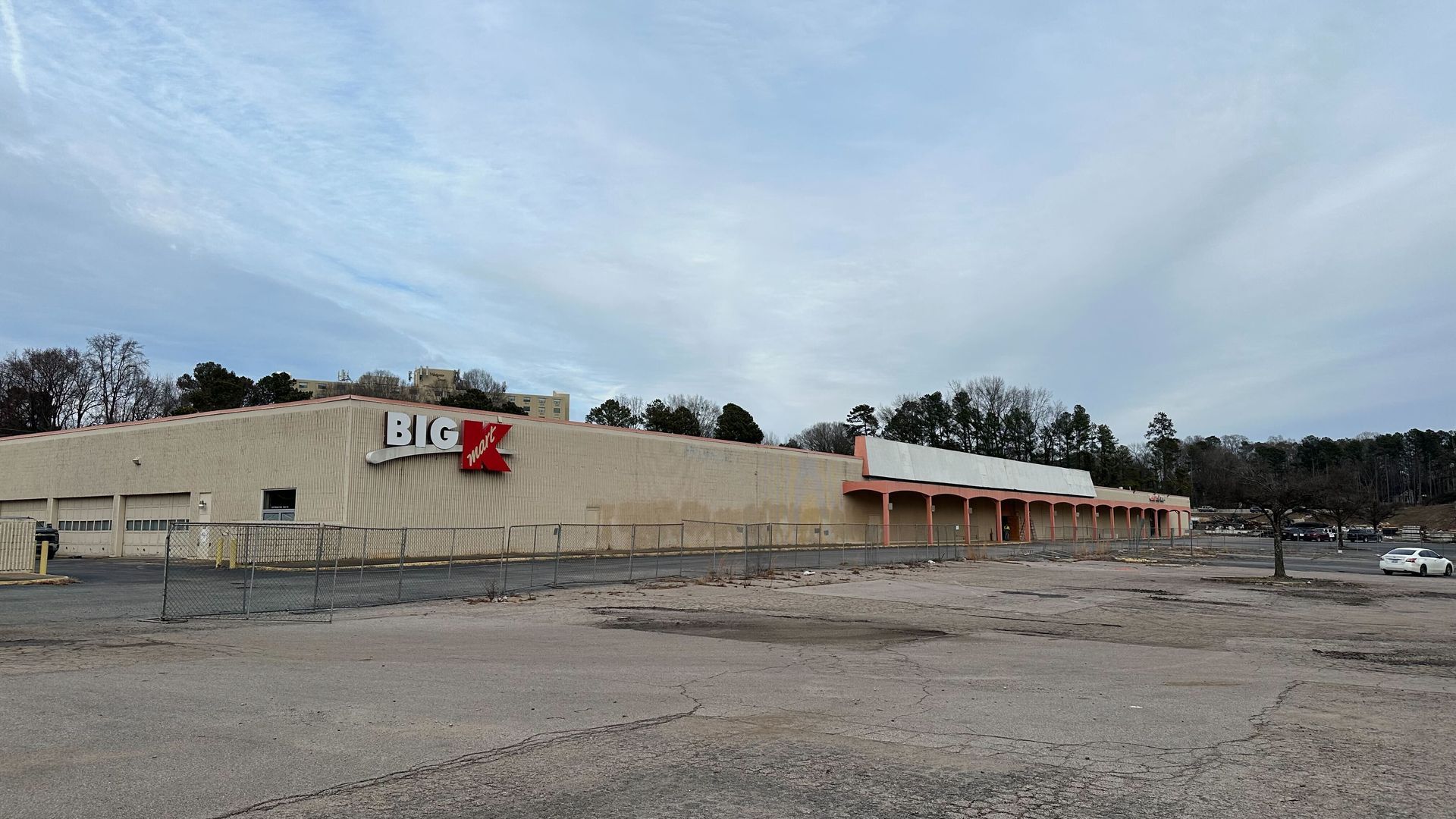 Large beige Big K Mart building with faded sign, enclosed by chain-link fence, empty cracked parking lot, cloudy sky, trees, and a few distant cars.