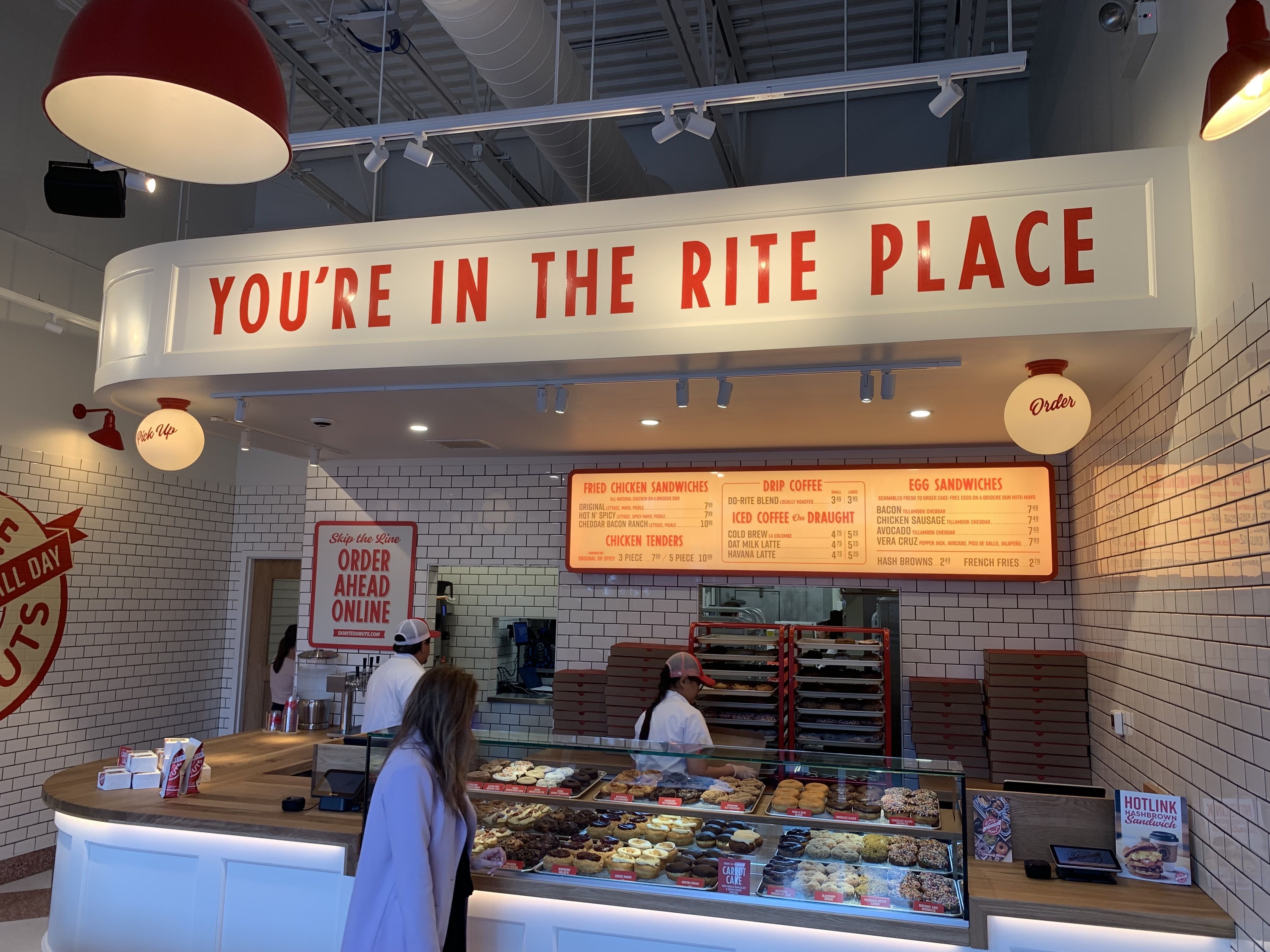 Bright cafe with curved white counter and glass pastry case. A bold sign reads "YOU'RE IN THE RITE PLACE" above orange menu boards; white tiles and warm lighting, with staff behind the counter.