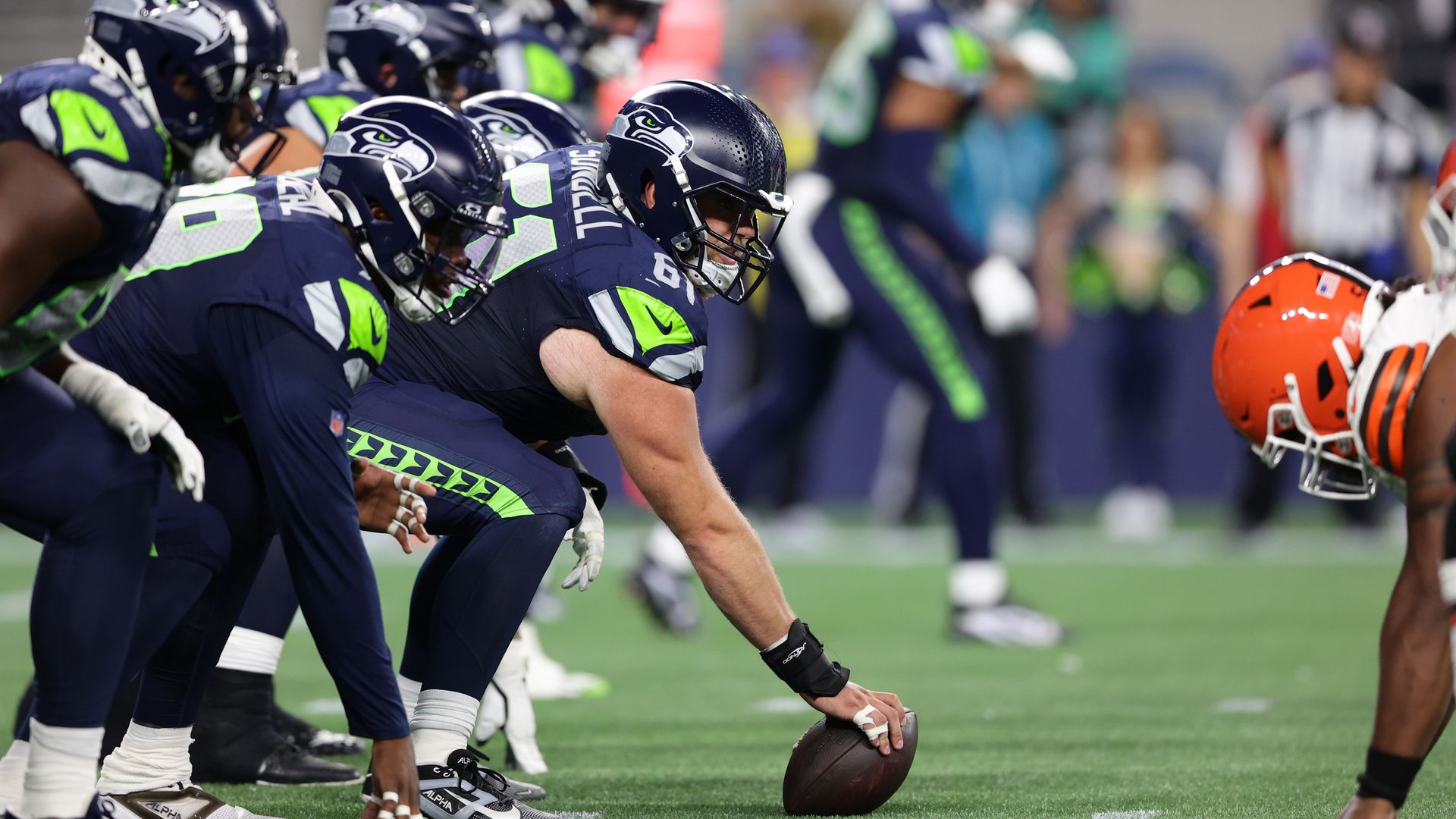 Seahawks players crouch at the line of scrimmage, with one holding a football, across from a player in an orange helmet.