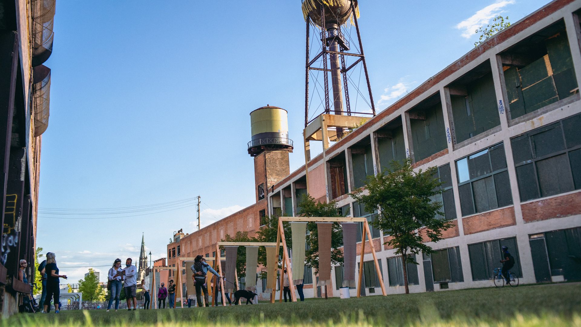 Urban scene with a tall metal water tower on a steel framework beside a brick building under a clear blue sky. People gather near a wooden-frame installation on the grassy area.