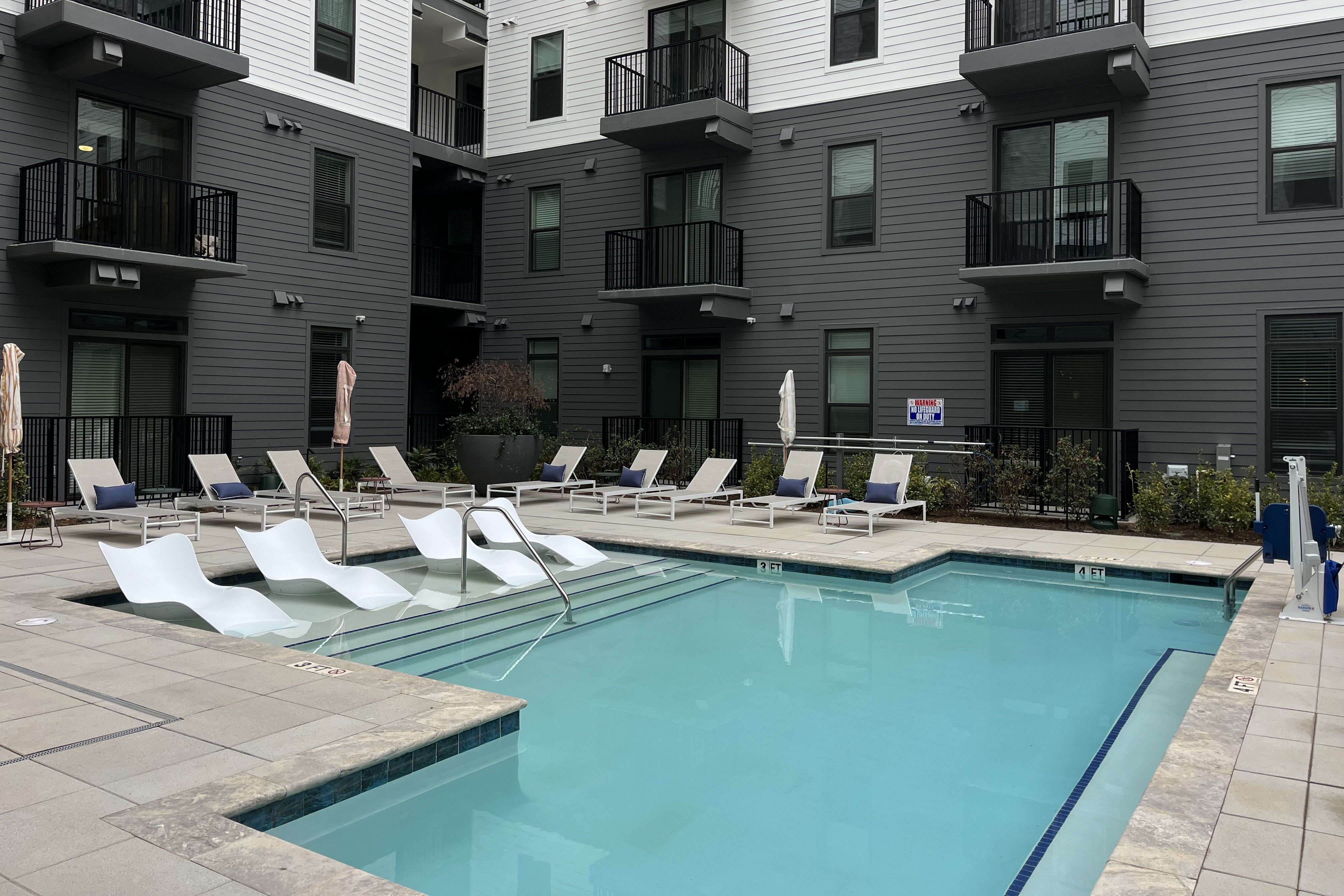 Modern apartment courtyard with a clean blue swimming pool, white lounge chairs with blue pillows, and gray building walls with black balconies in the background.