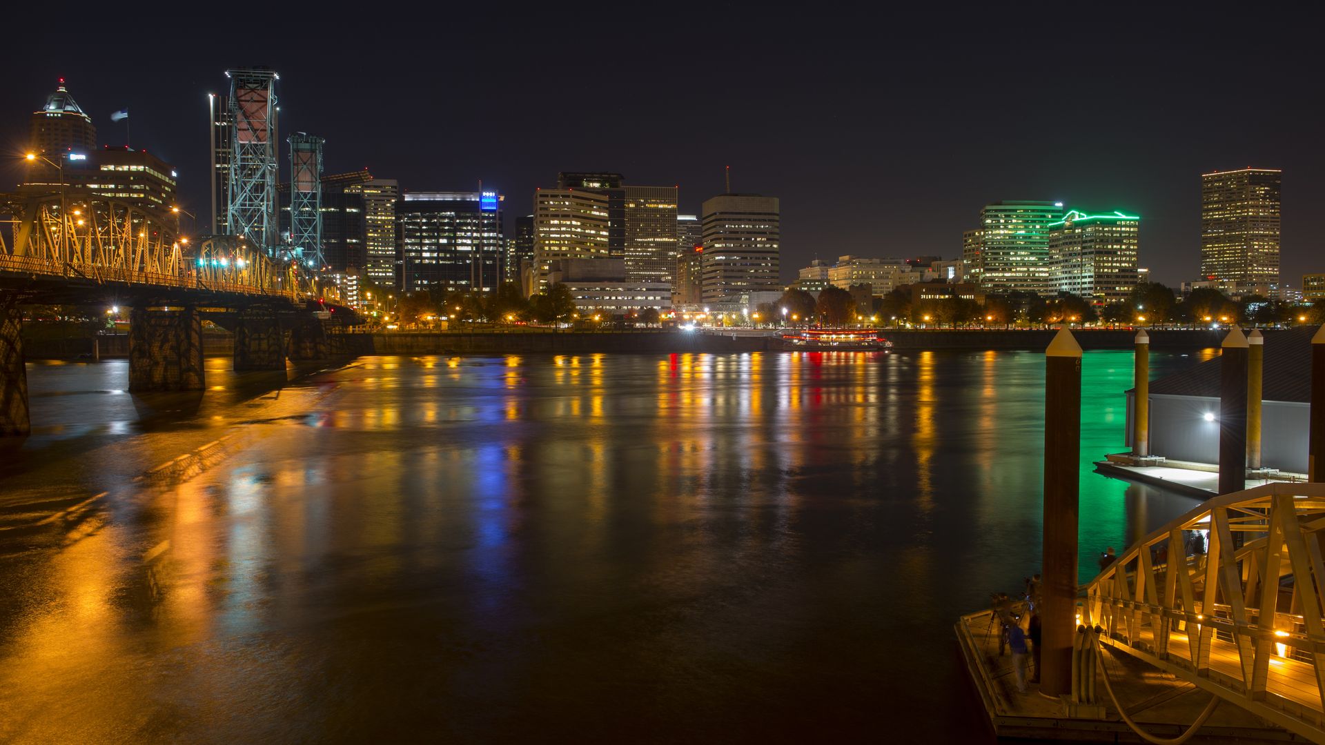 Night city skyline over a river with a lit left bridge, tall center buildings, and green neon towers on the right; colorful reflections ripple on the water as a pier extends in the foreground.
