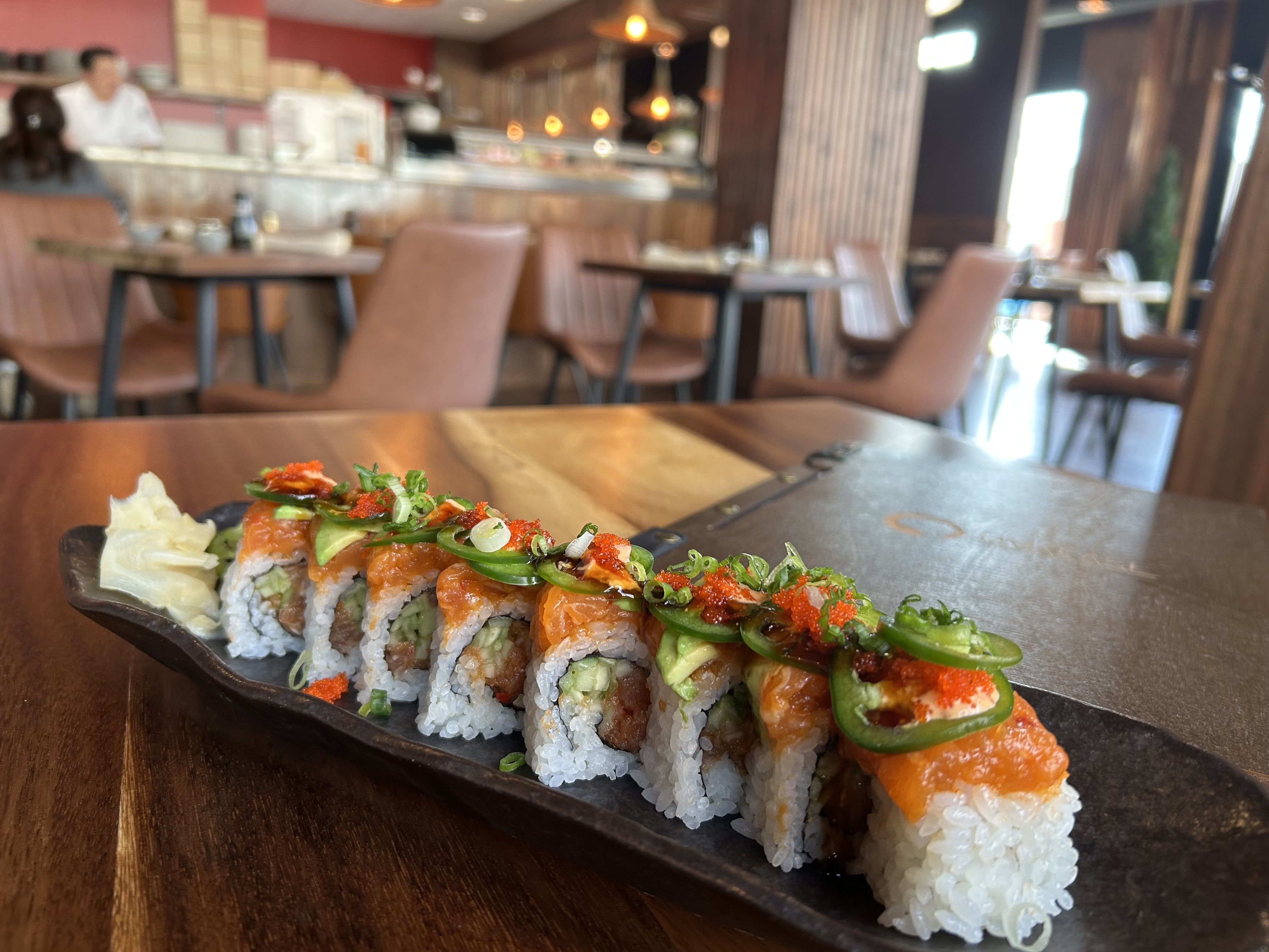 Plate of sushi rolls topped with spicy salmon, jalapeño slices, green onions, and red roe on a wooden table in a sushi restaurant with brown chairs and a sushi bar in the background.