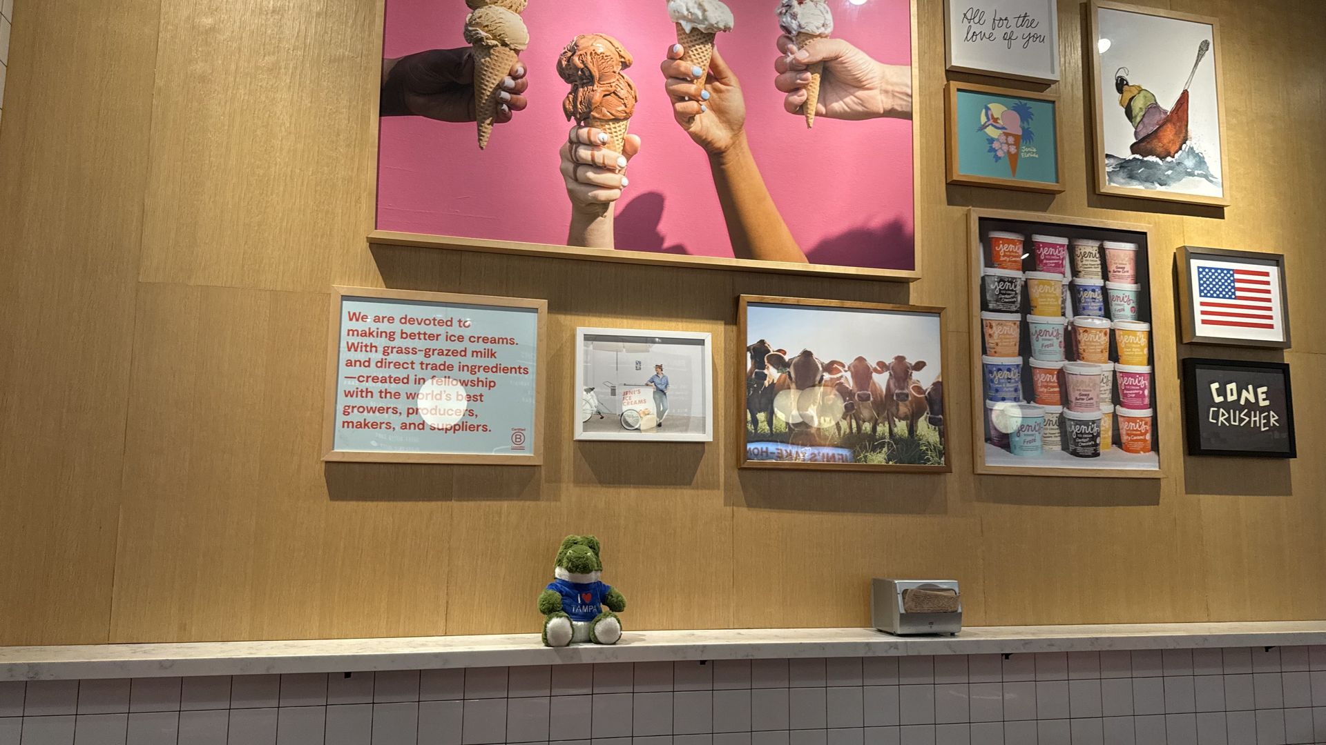 Wall display with a large pink photo of hands holding ice cream cones, surrounded by framed art including ice cream cans and a U.S. flag, plus a small green teddy bear in a blue shirt on the counter.