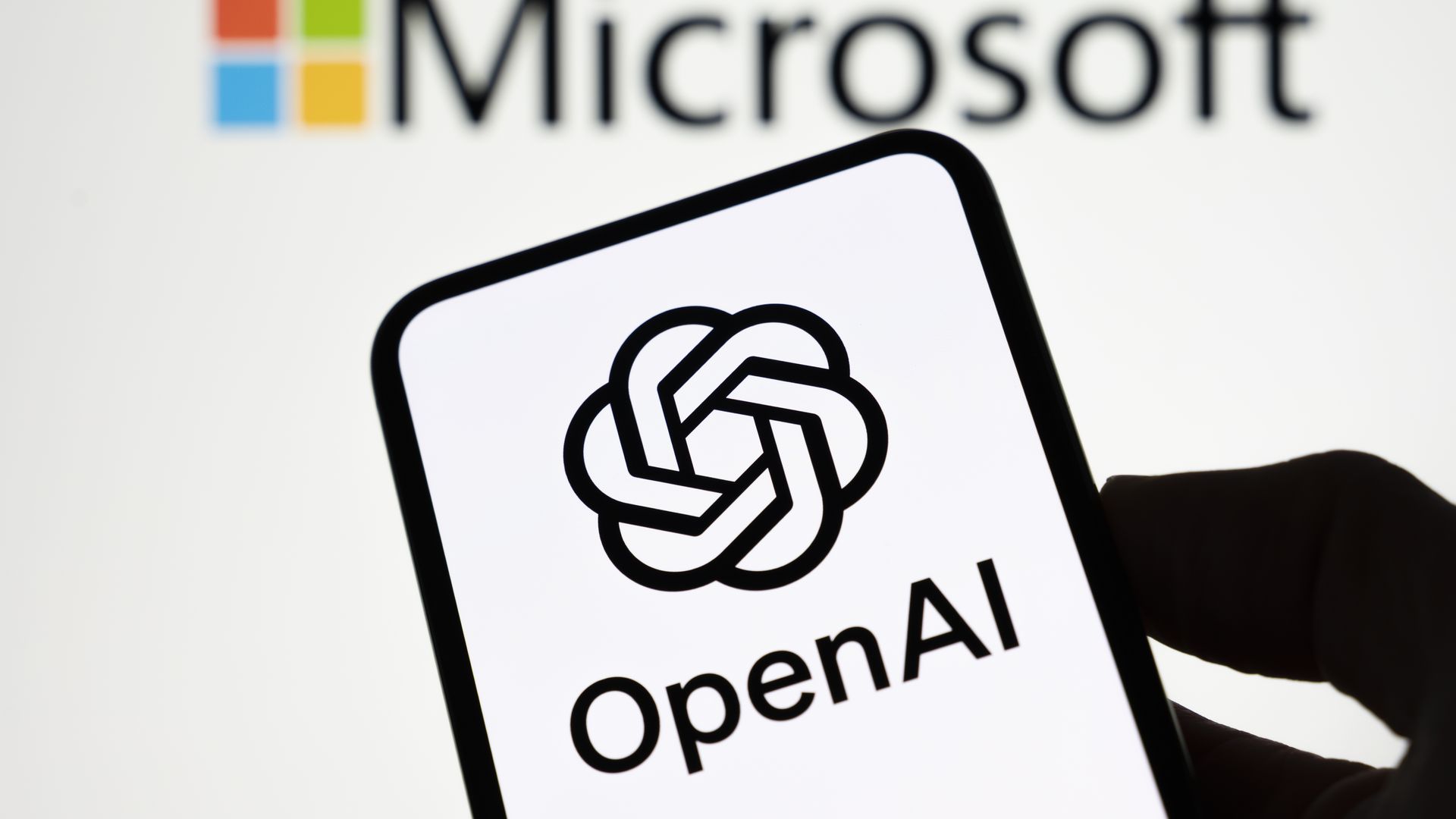 Close-up of a hand holding a smartphone displaying the OpenAI logo (a black knot) and the word OpenAI, with a blurred Microsoft logo in the background.