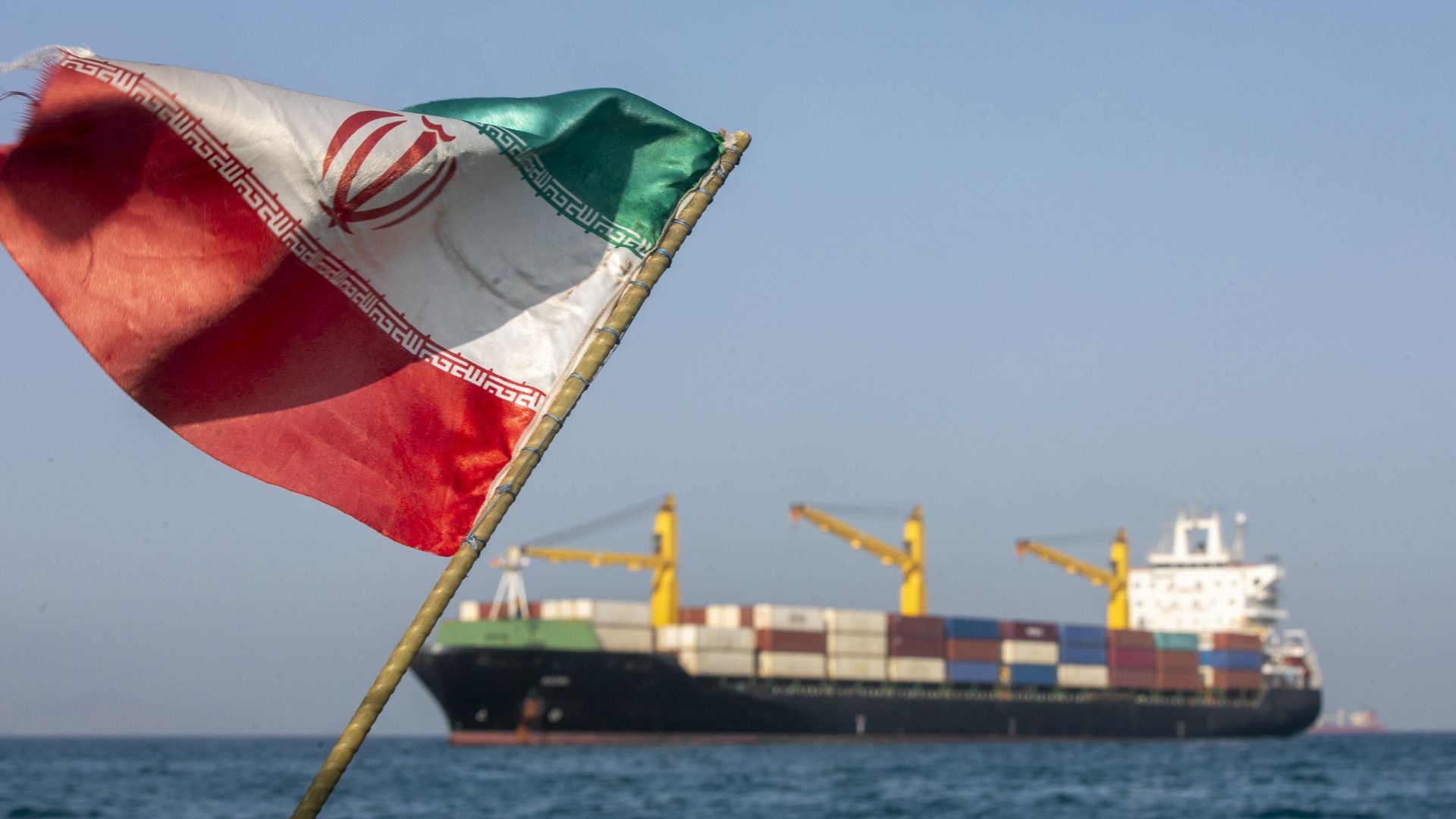 An Iranian flag flies in the foreground as a cargo ship sits in the Strait of Hormuz near Larak Island.