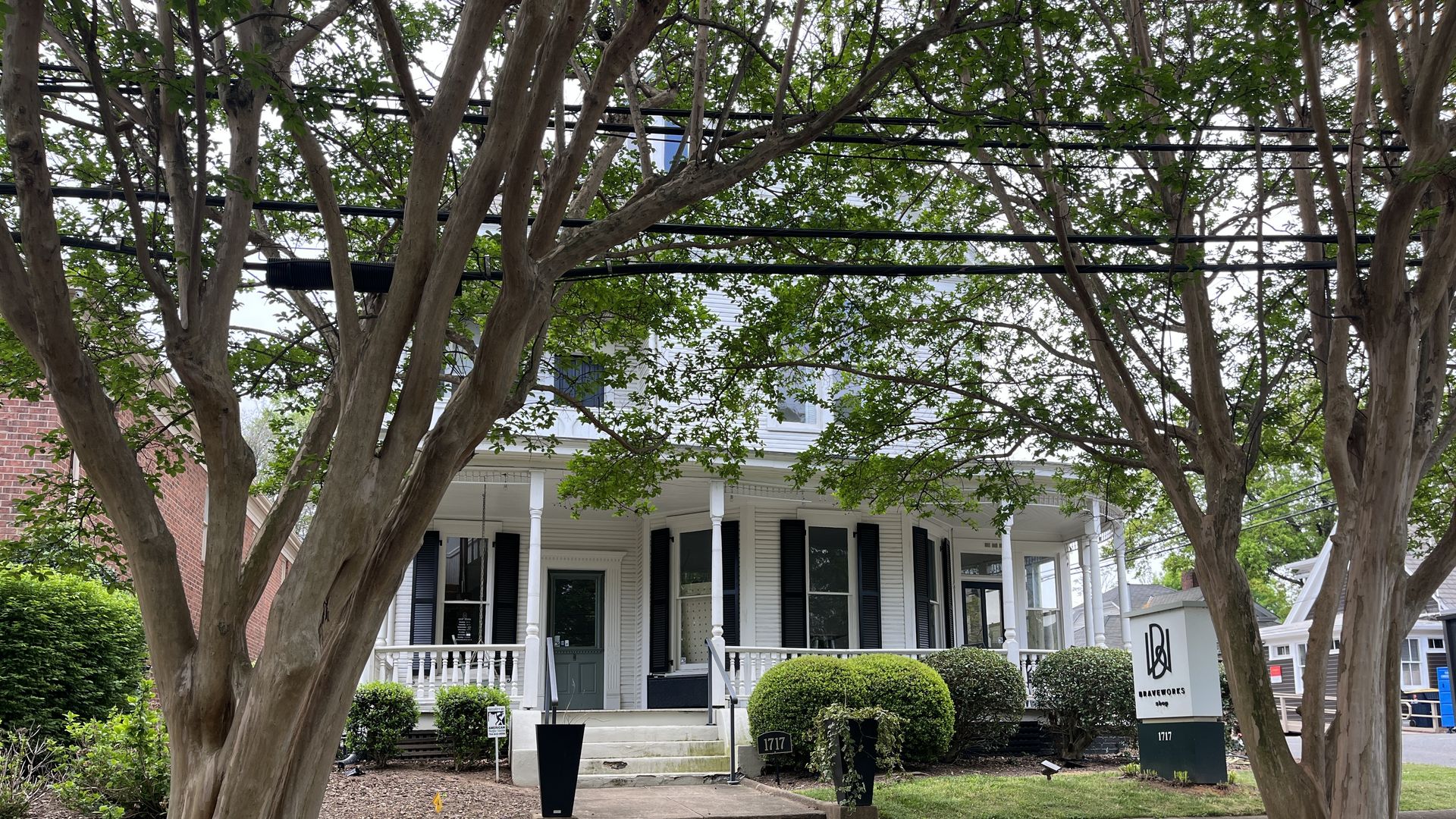 White house with a wraparound porch, framed by two large trees. Green front door, black shutters, trimmed round shrubs, and a sidewalk with a sign in the yard.