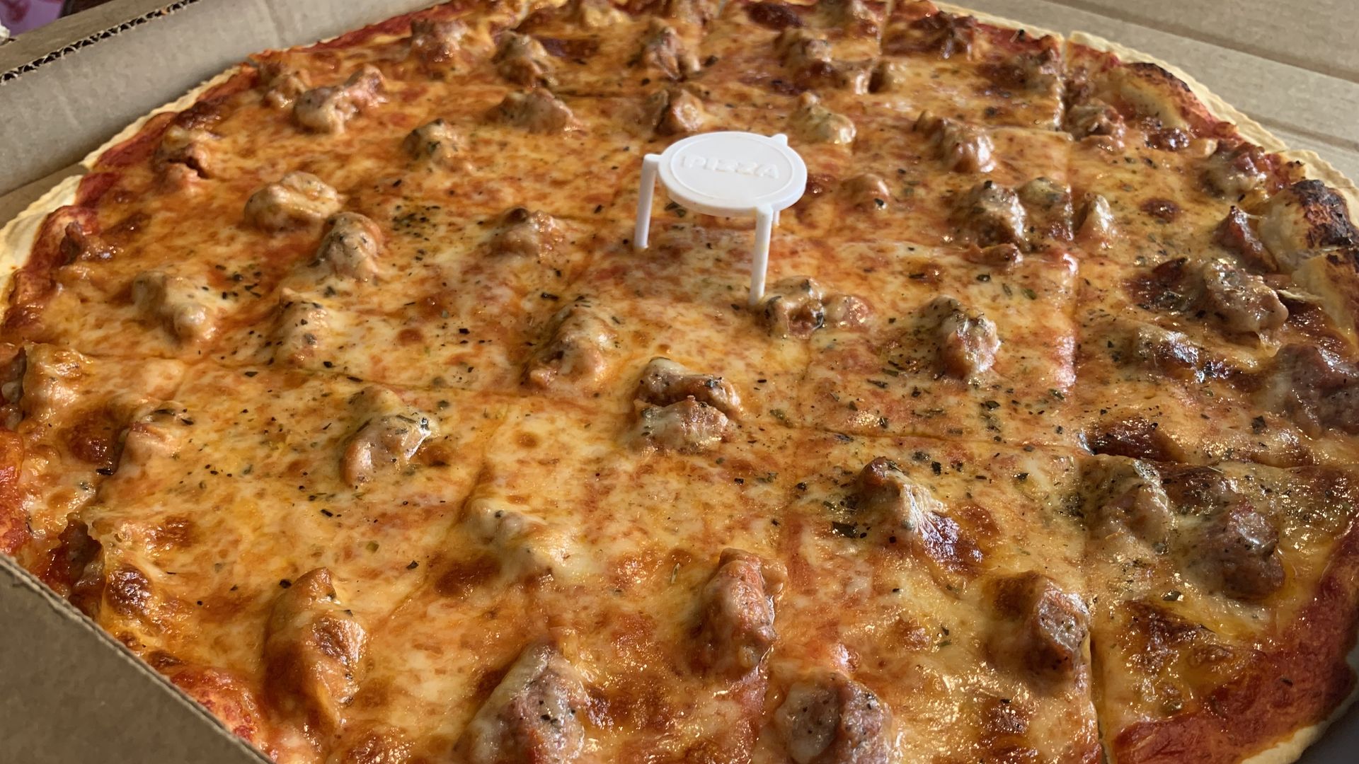 Large cheese and sausage pizza in an open box, with bubbly melted cheese, herbs, and browned crust. A white plastic pizza saver stands in the center.