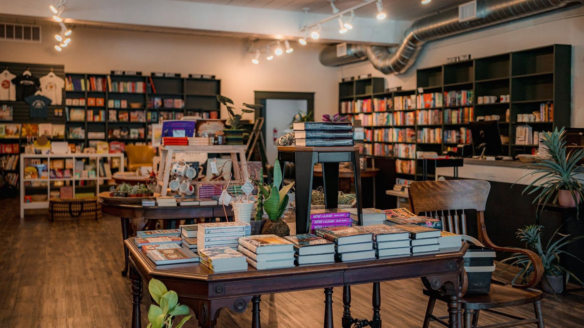 Cozy bookstore interior with dark shelves lining the walls, stacks of books on a central table, warm string lights, wooden floors, and potted plants; a large silver duct runs along the ceiling.