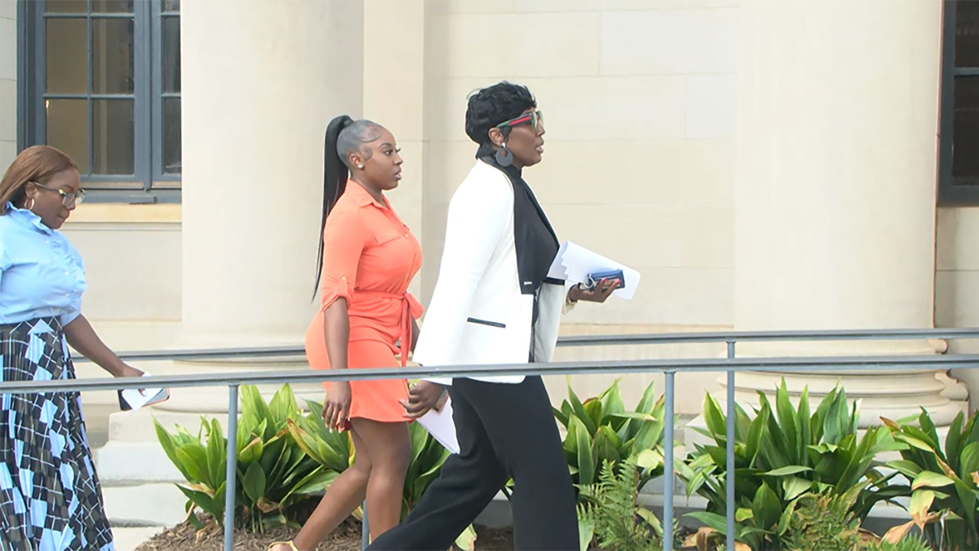 Three women walk along a campus walkway beside a beige columned building. Left: light-blue blouse and patterned skirt; center: orange dress; right: white blazer with black outfit, holding papers.