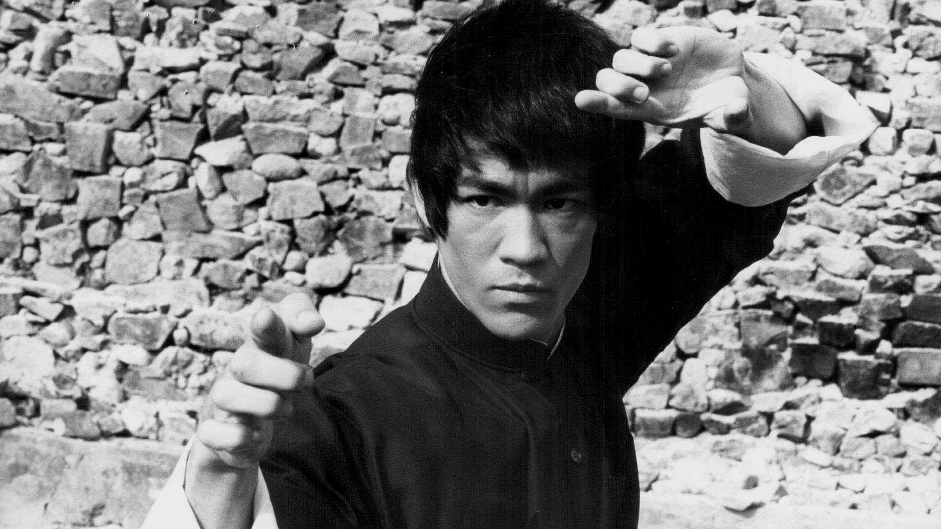Bruce Lee stands with his arms up as if ready to engage in a fight. Photo is black and white.