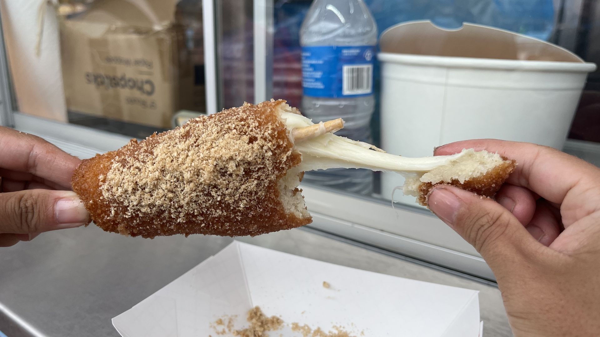Two hands pull apart a breaded, fried mozzarella stick, cheese stretching between halves. In the background a blue water bottle and a white takeout cup sit by a window.
