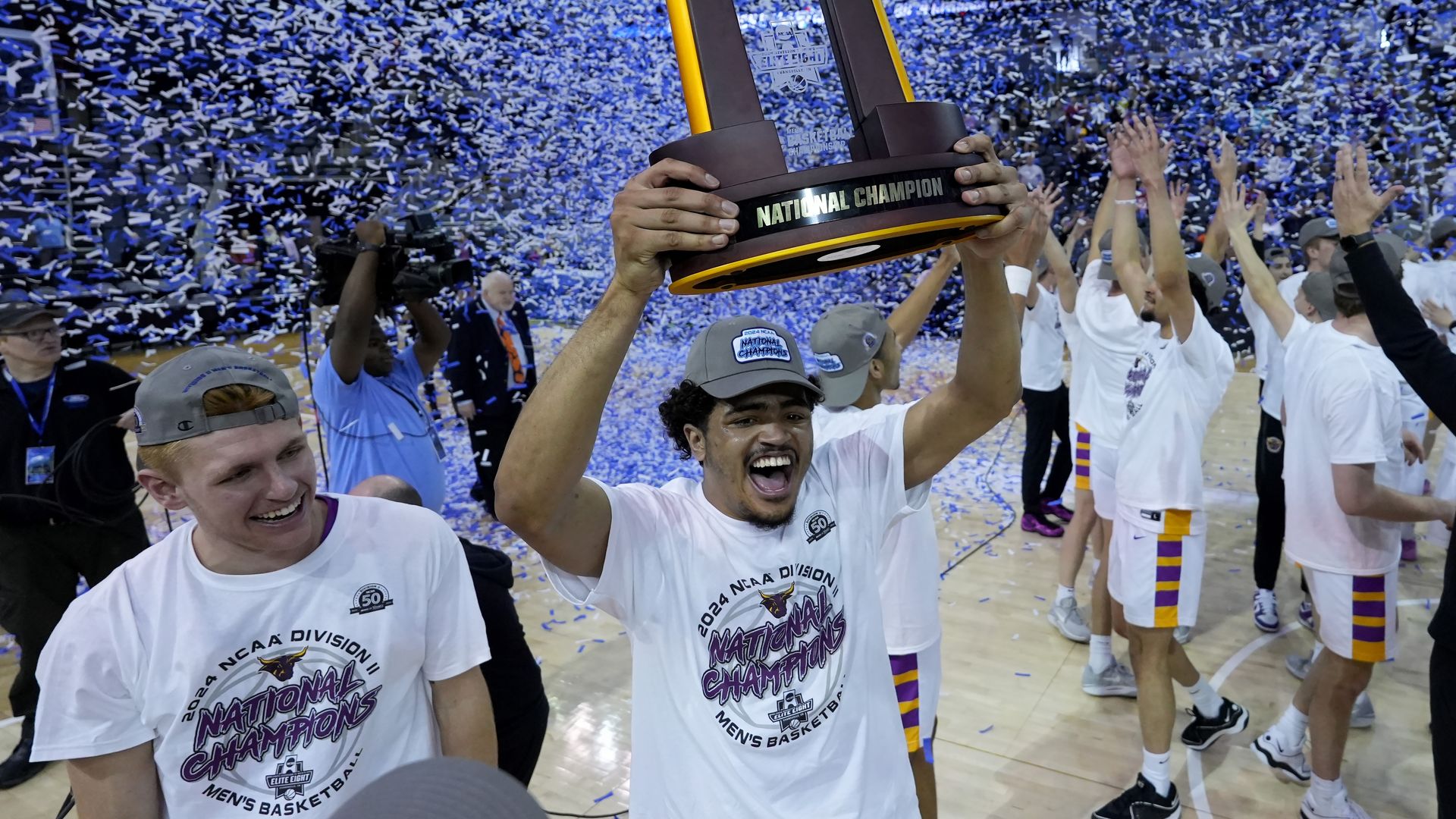 A man with brown skin shouts with joy and holds up a trophy that says "National Championship" as blue confetti sprays behind him.