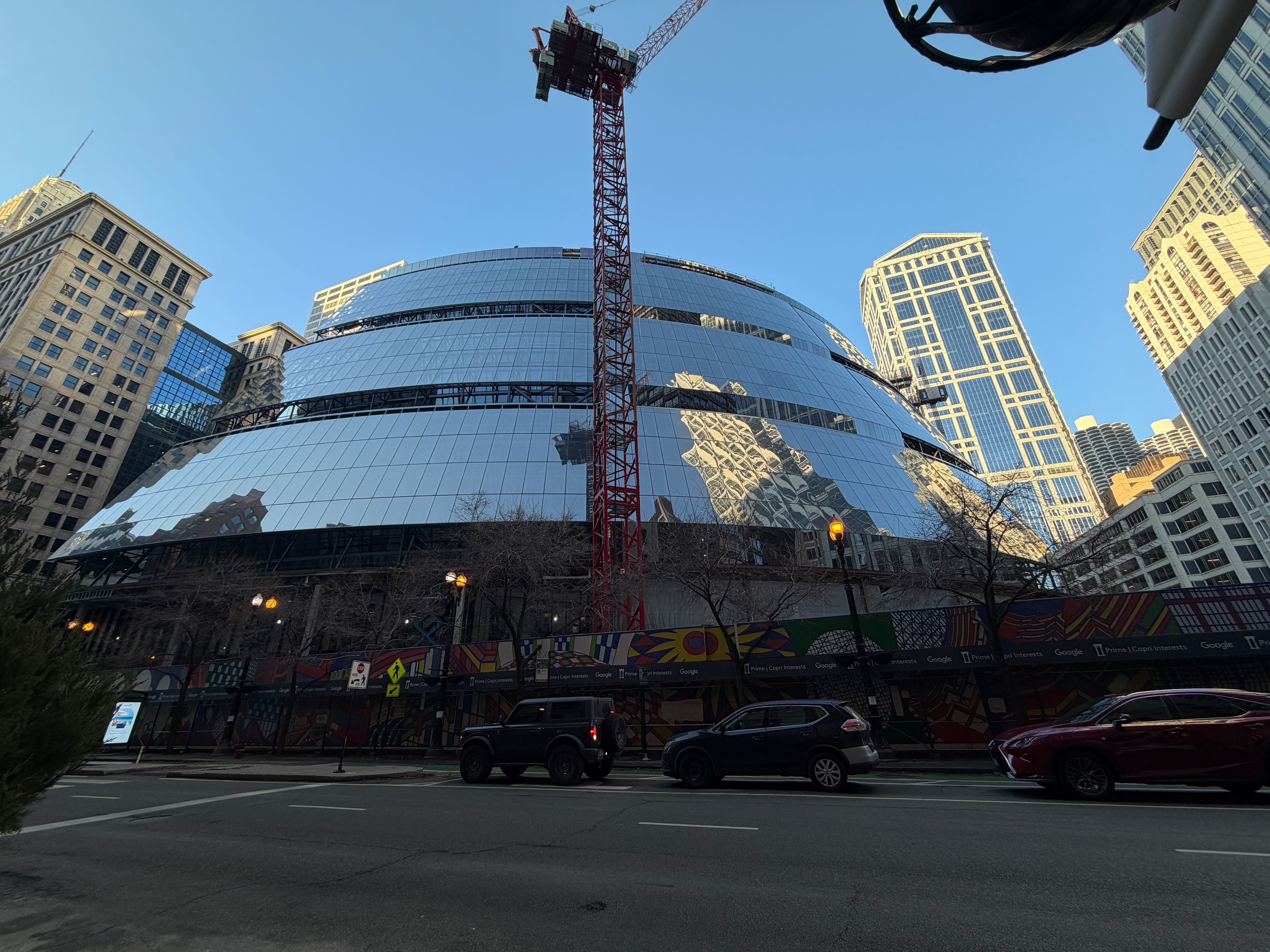 Curved glass building under construction with a tall red crane, surrounded by tall skyscrapers, cars parked on the street, and colorful construction barriers under a clear blue sky.