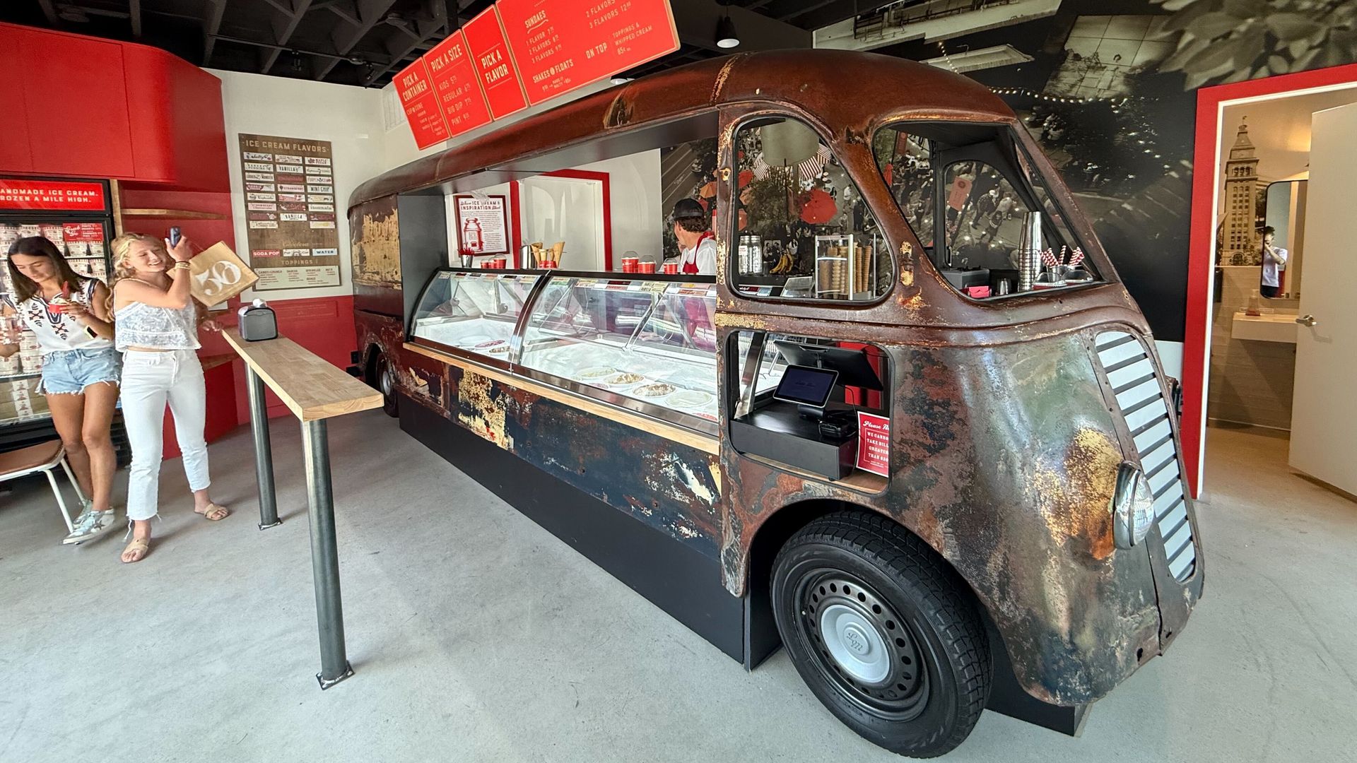 Interior of an ice cream shop with a vintage, rusty van converted into an ice cream display counter, red menu boards above, and two women near a wooden table on the left.