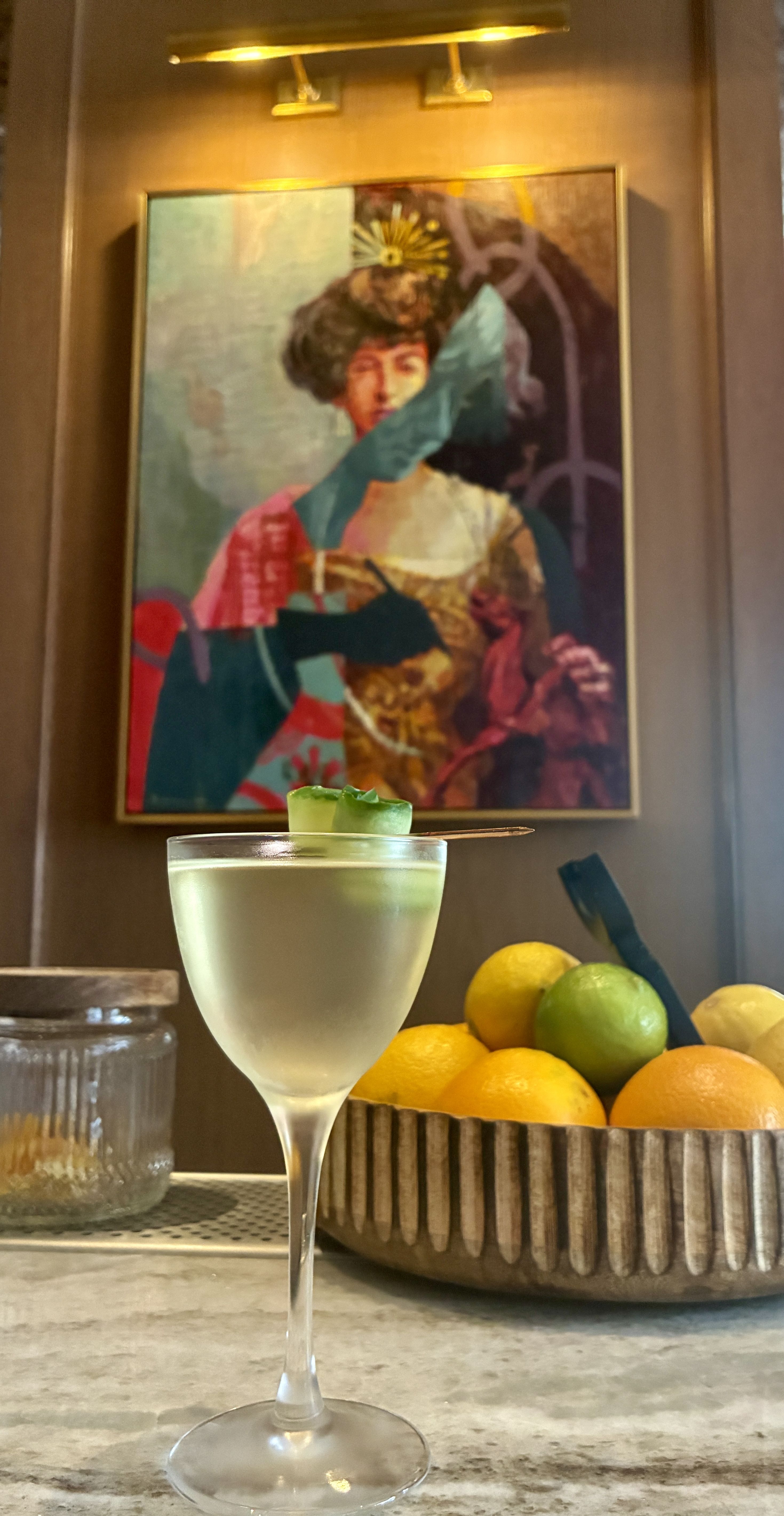 A pale cocktail in a stemmed glass with a lime garnish sits on a marble counter. Behind it, a colorful framed painting of a crowned woman hangs, with a bowl of lemons and limes nearby.