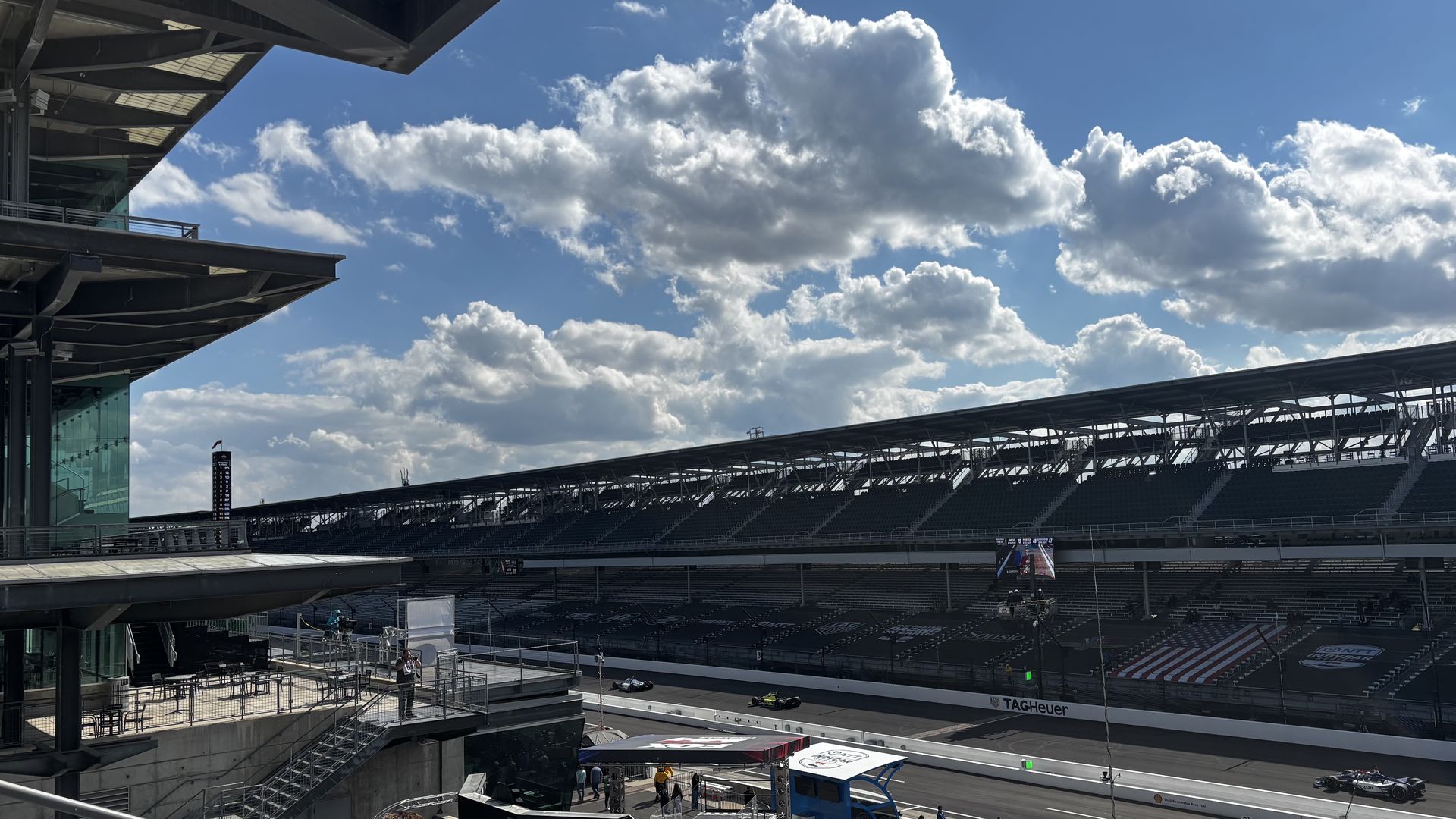 Wide view of a race track stadium: steel and glass structures on the left, empty grandstands on the right, a race car in the pit lane, and a bright blue sky with fluffy white clouds.