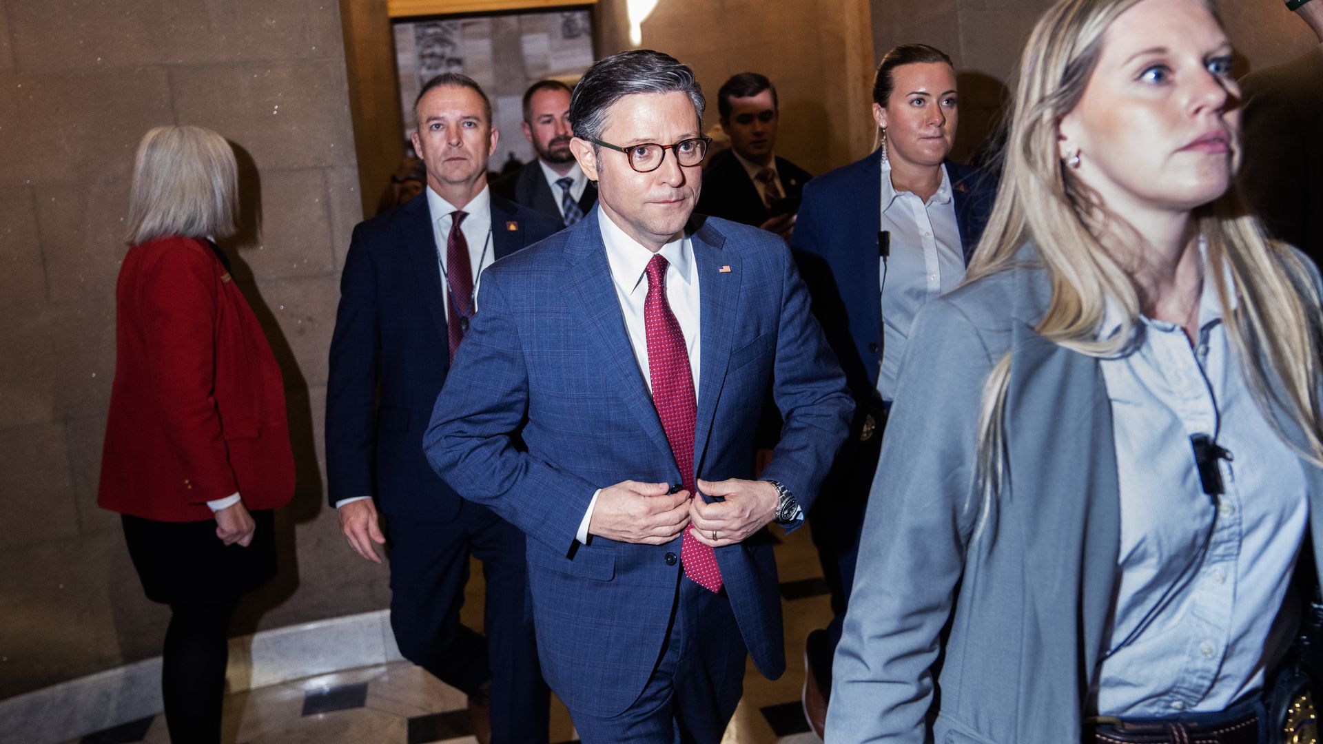 House Speaker Mike Johnson, wearing a blue suit, white shirt and red tie, walking through the Capitol with his security detail.