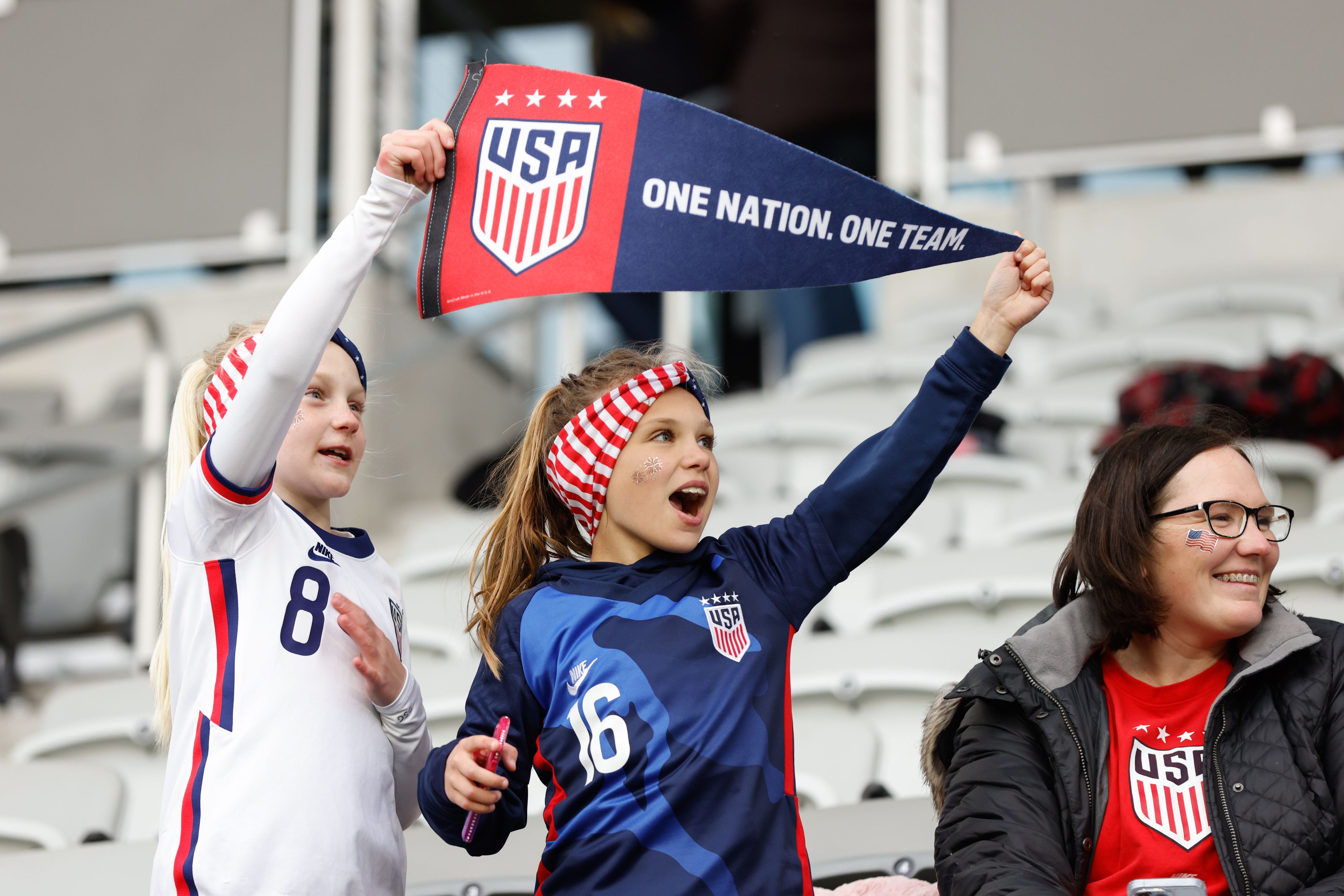 Young fans of the USWNT soccer team hold a pennant reading "USA, One Nation. One Team."