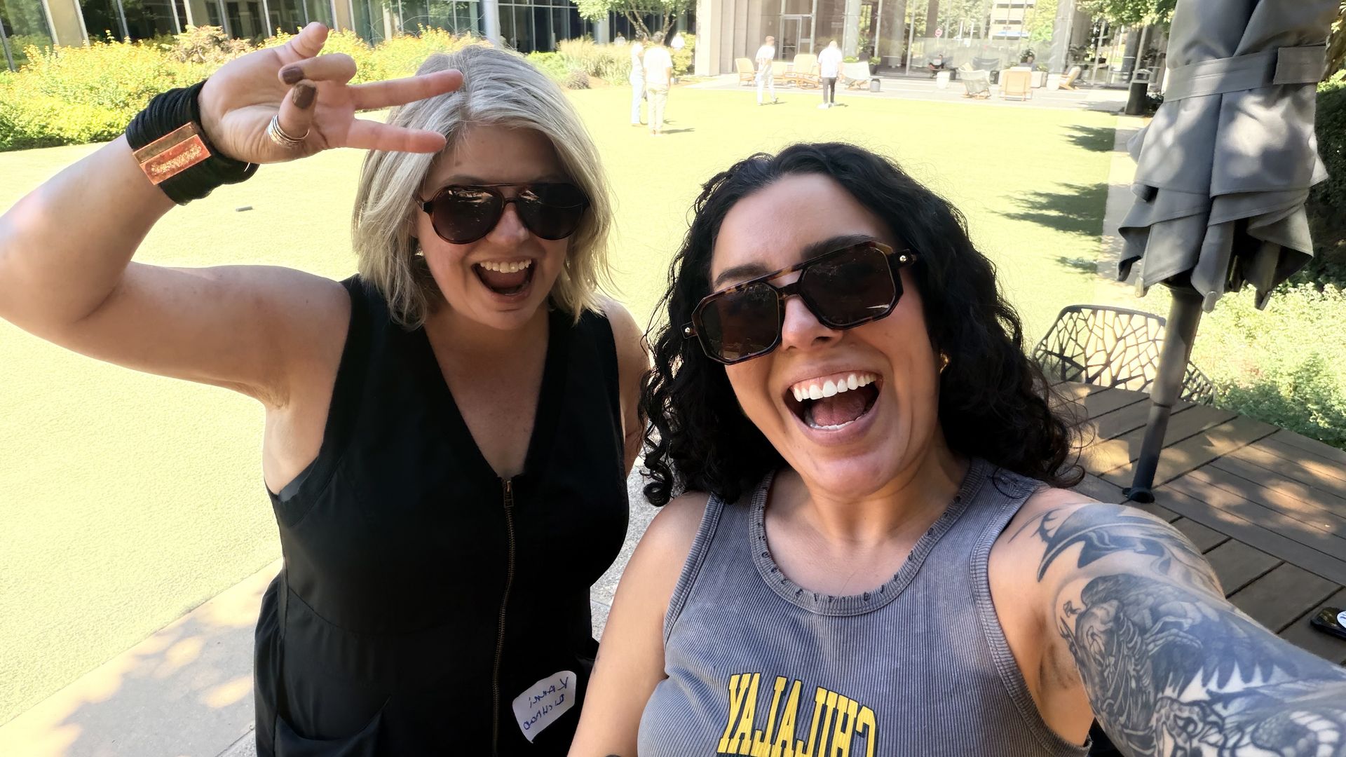 Two women wearing sunglasses take a sunny outdoor selfie. Left blond makes a peace sign; right dark-haired wears a gray tank top as they smile. Green lawn and glass building in the background.