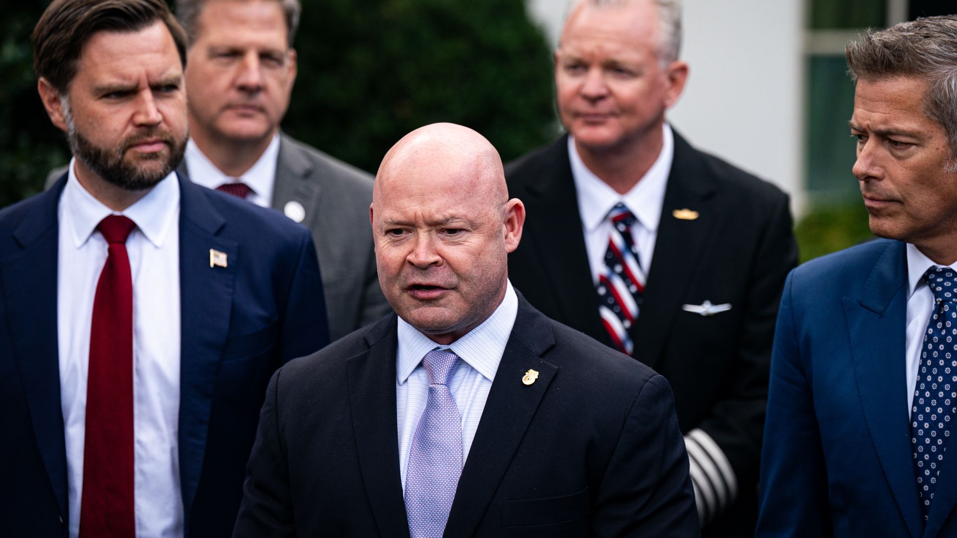 Teamsters president Sean O'Brien, wearing a dark blue suit, is flanked by Vice President J.D. Vance and Transportation Secretary Sean Duffy and two other men, all wearing suits, outside a white building with hedges in front of it.