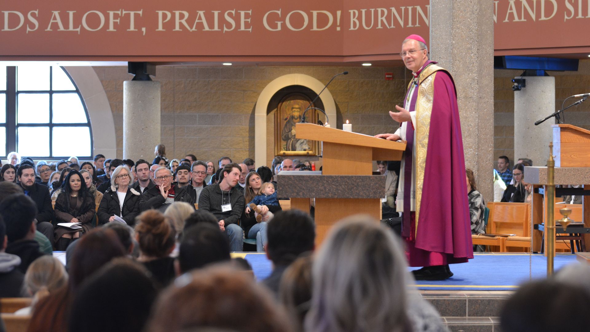 Magenta-robed bishop speaks from a wooden lectern to a seated congregation in a church; behind him, a candle, an icon, and a large banner reading PRAISE GOD.