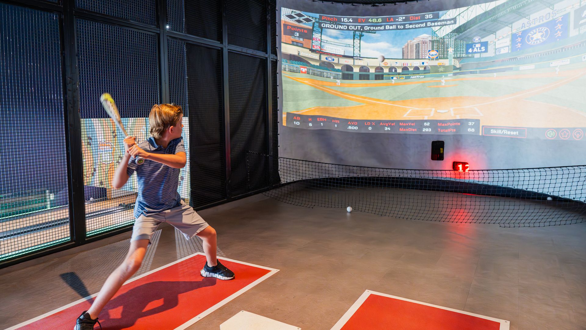 Young boy in a batting cage swings a bat toward a large curved screen showing a baseball stadium; dark netting frames the scene, red mats under the plate, a ball on the ground nearby.