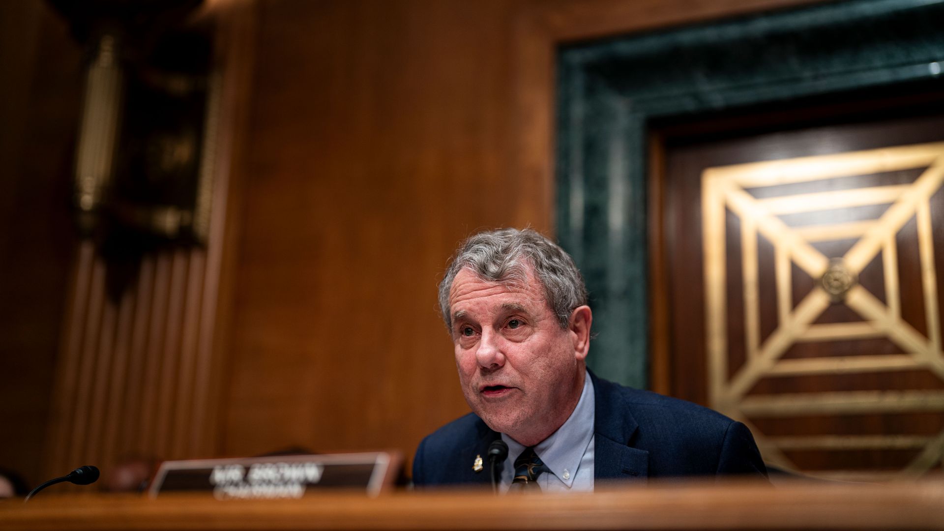 A man with gray hair wearing a dark blue suit and patterned tie speaks into a microphone in a wood-paneled room with a decorative wall and plaque in the background.