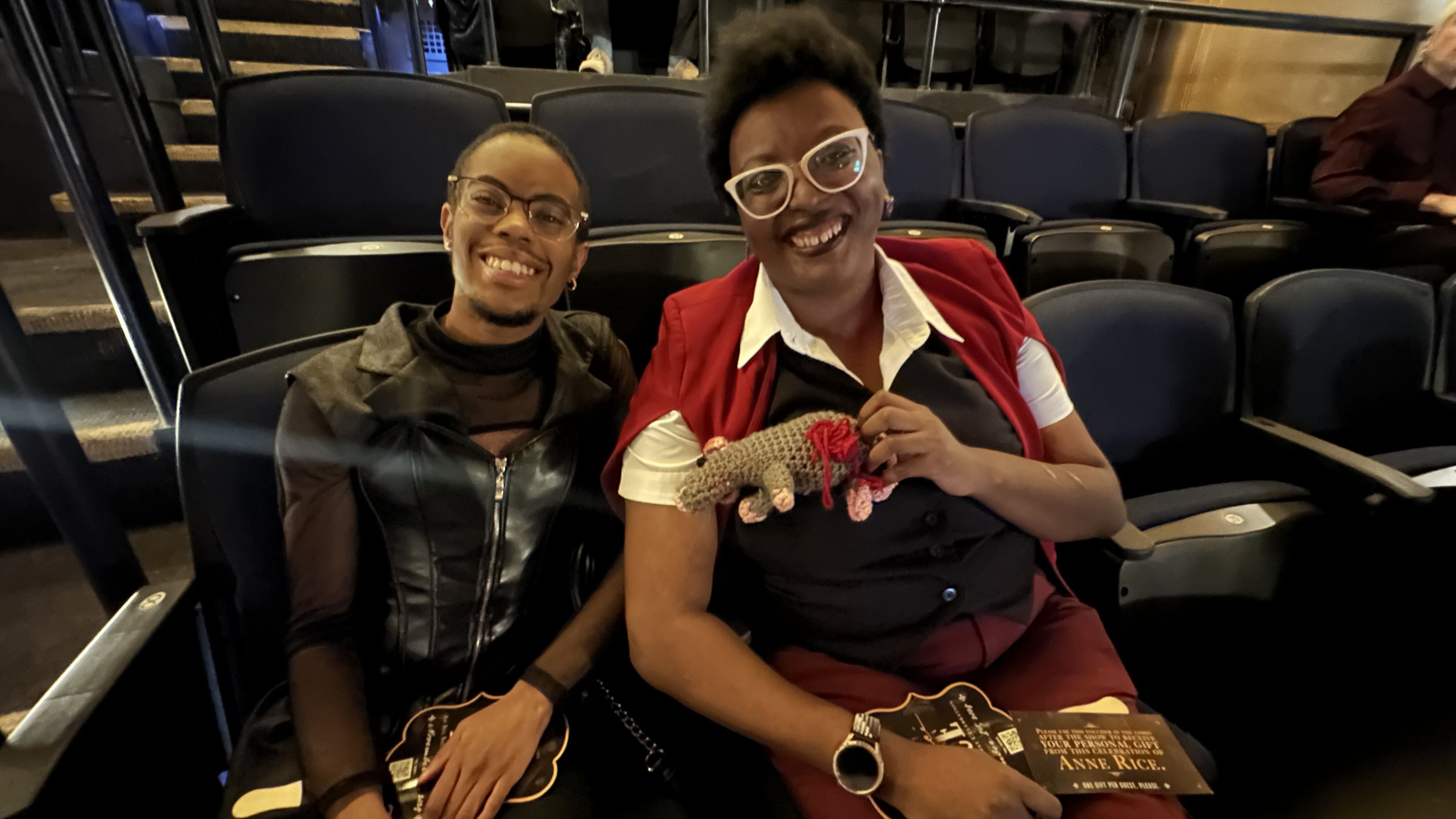 Two smiling people sitting in dark blue theater seats, one wearing glasses and a black outfit, the other with white glasses, a red vest, and holding a crocheted animal.