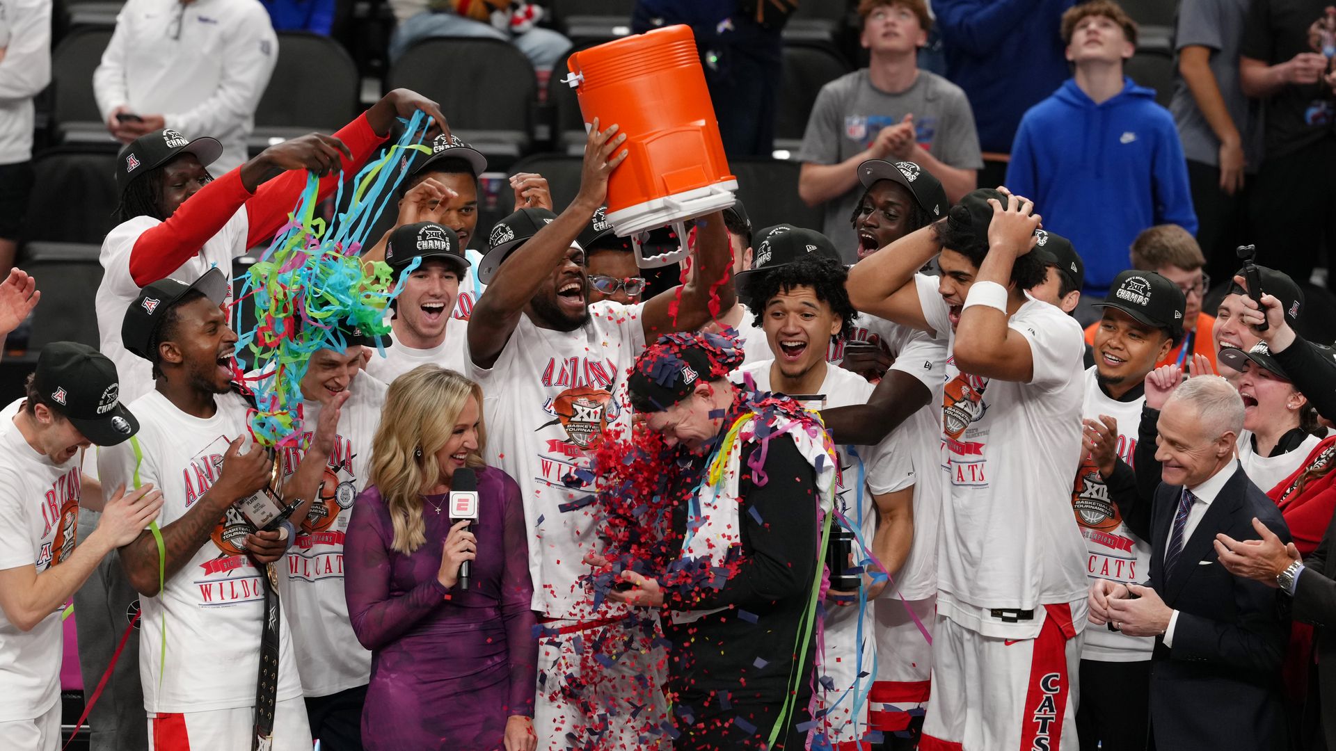 A group of men's college basketball players in white and red shirts, white t-shirts and black ballcaps celebrate while a player dumps confetti from an orange cooler on a coach wearing all black, with a woman wearing purple and holding a microphone standing next to him. 