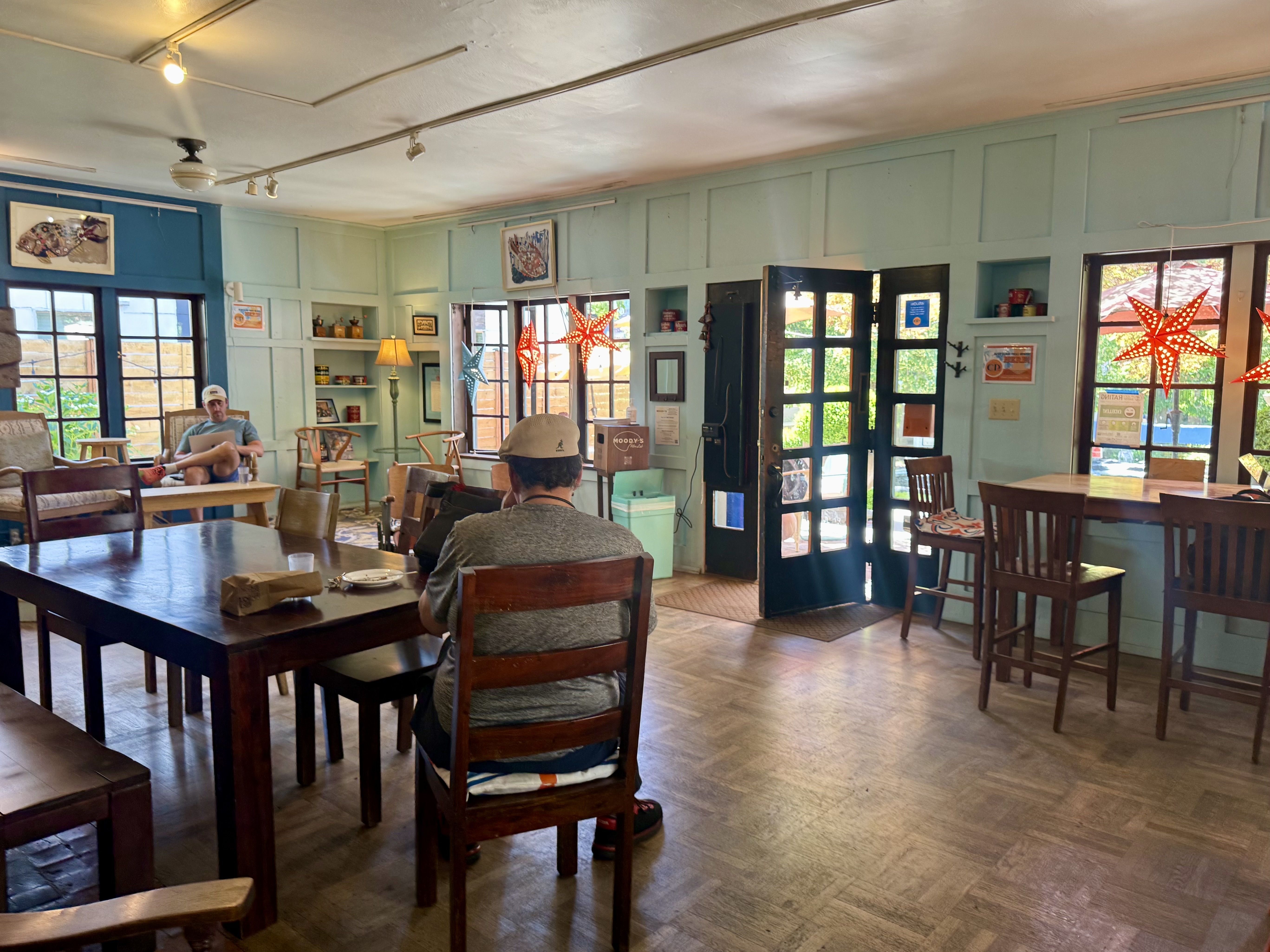 Bright cafe interior with pastel blue walls, wooden tables and chairs, star-shaped red and blue lanterns by windows and doors, and a man sitting with a laptop.