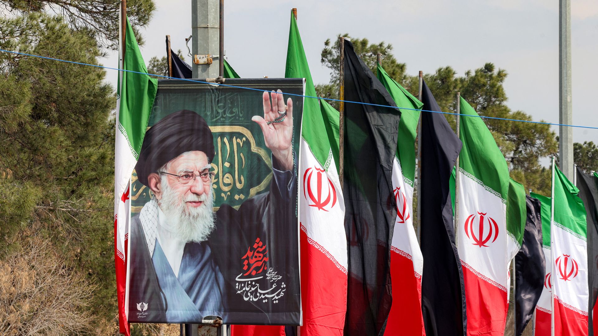 Row of roadside banners featuring a large poster of a bearded cleric raising his hand; green, white, and red Iranian flags flank the display with dark banners and trees in the background.