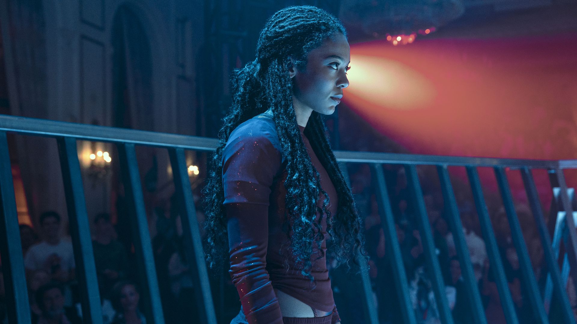 Side profile of a serious young woman with long curly hair in a dark red outfit, standing in a dimly lit venue with blue and red lighting and blurred audience behind a railing.