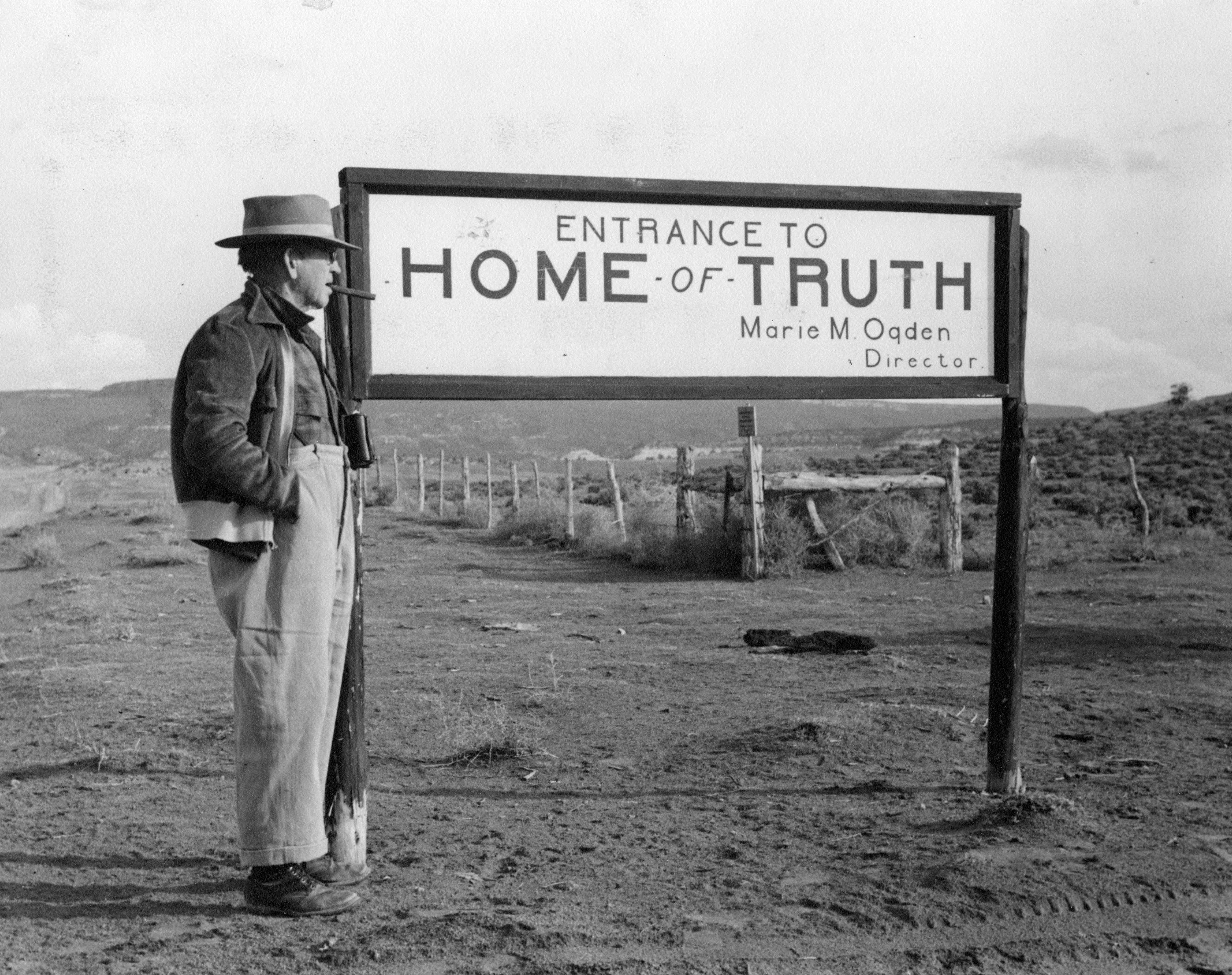 Black and white photo of a man standing next to a sign that reads "Entrance to Home of Truth"