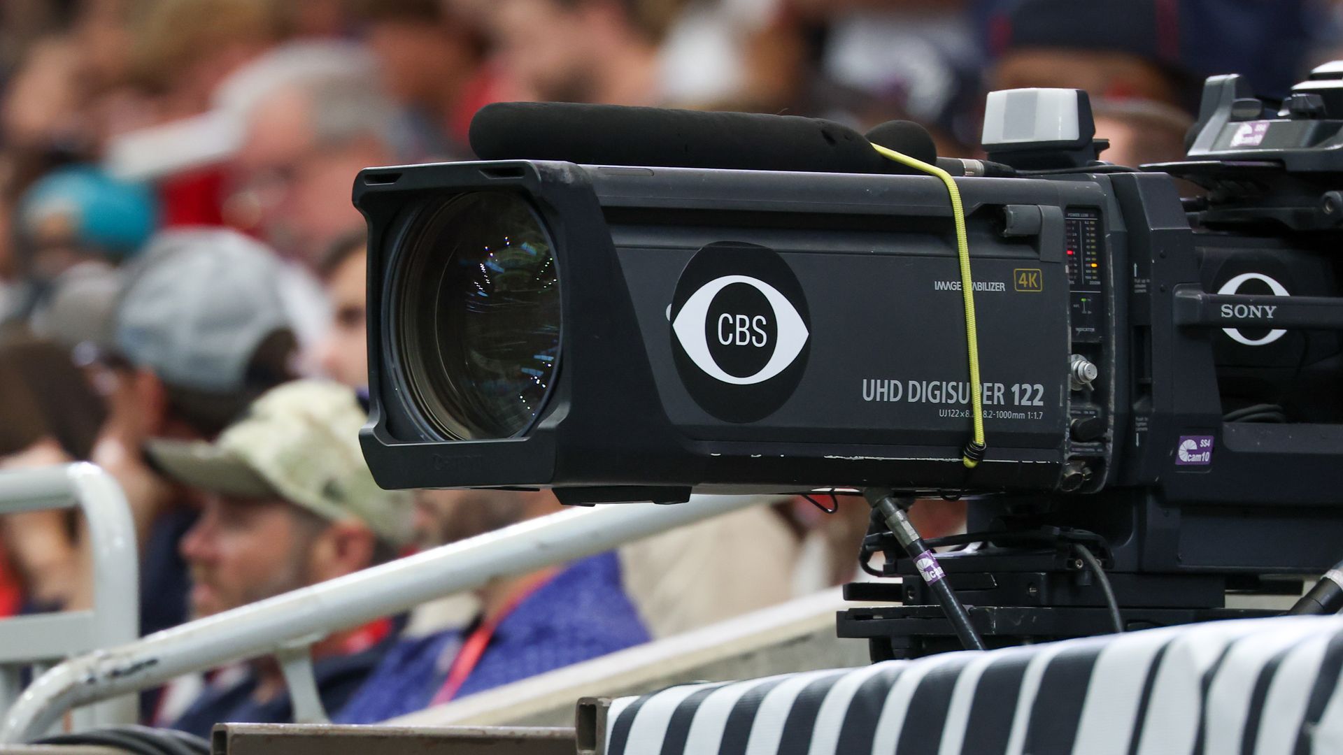Close-up of a broadcast camera with a CBS logo, yellow cable crossing the top, a white viewfinder, and a striped barrier in the foreground; blurred crowd in the background
